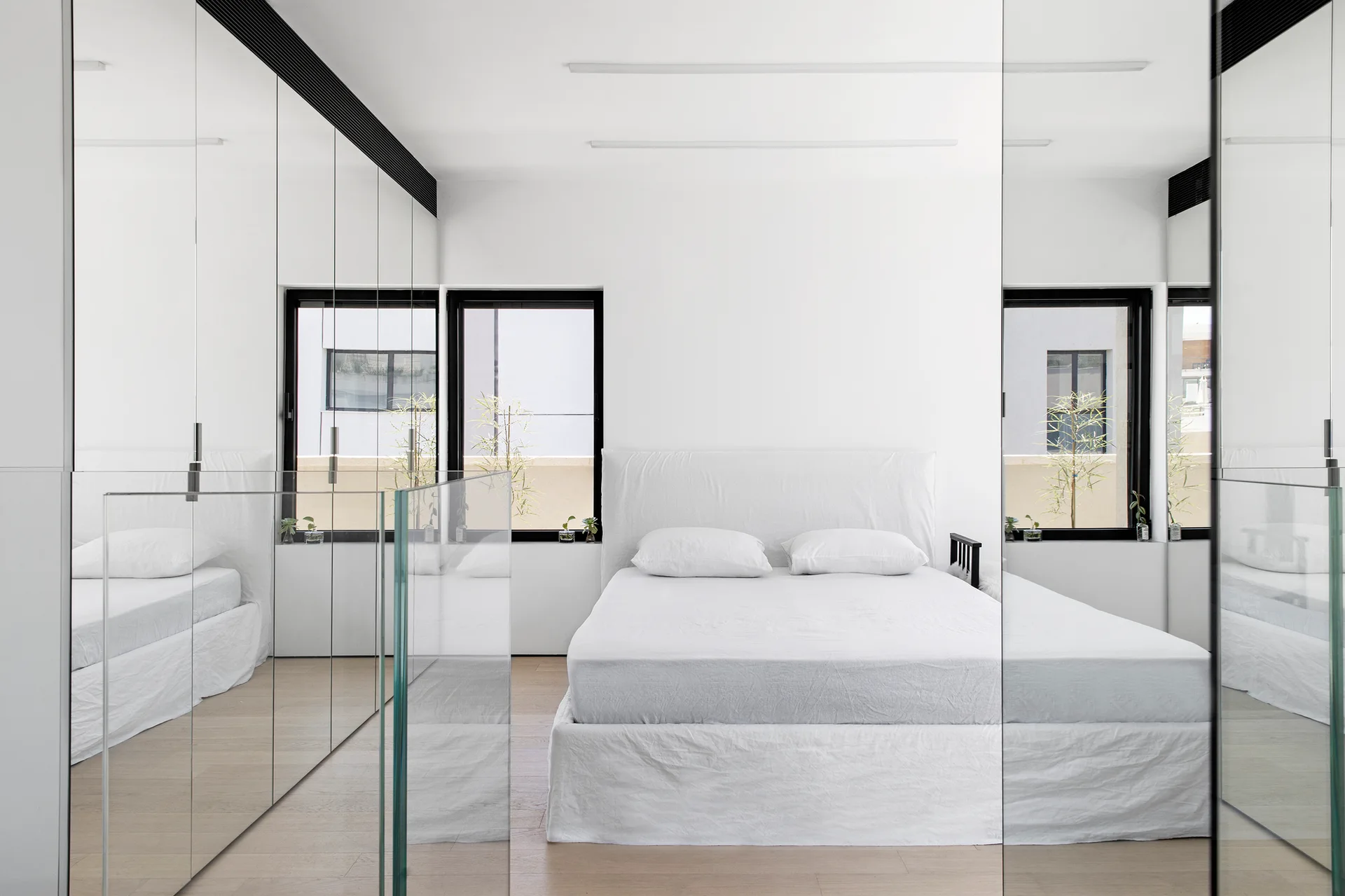A minimalist white bedroom features a bed with white bedding centered between large floor-to-ceiling mirrored wardrobe doors and black-framed windows.