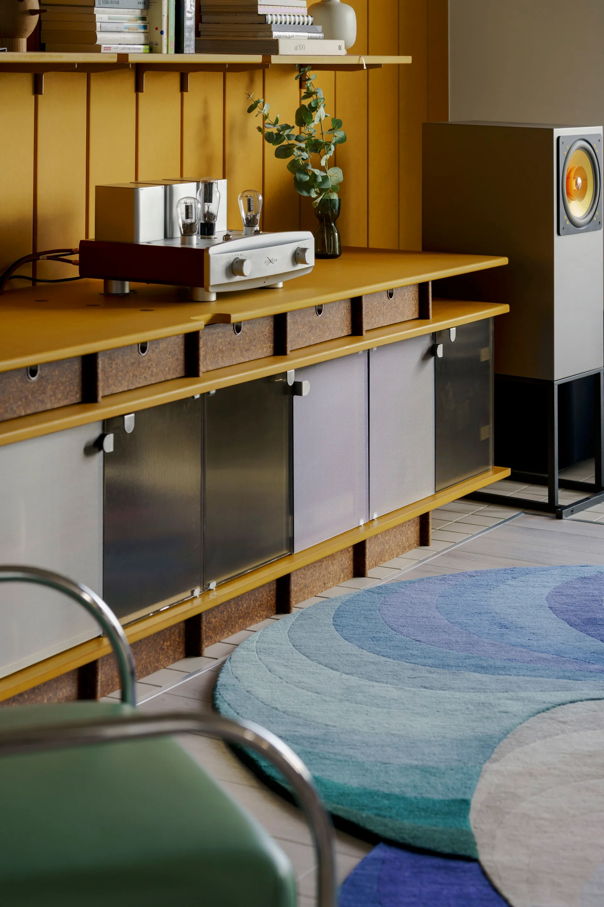 A custom yellow media console with a tube amplifier sits against a matching paneled wall beside a blue patterned rug and a tall speaker.