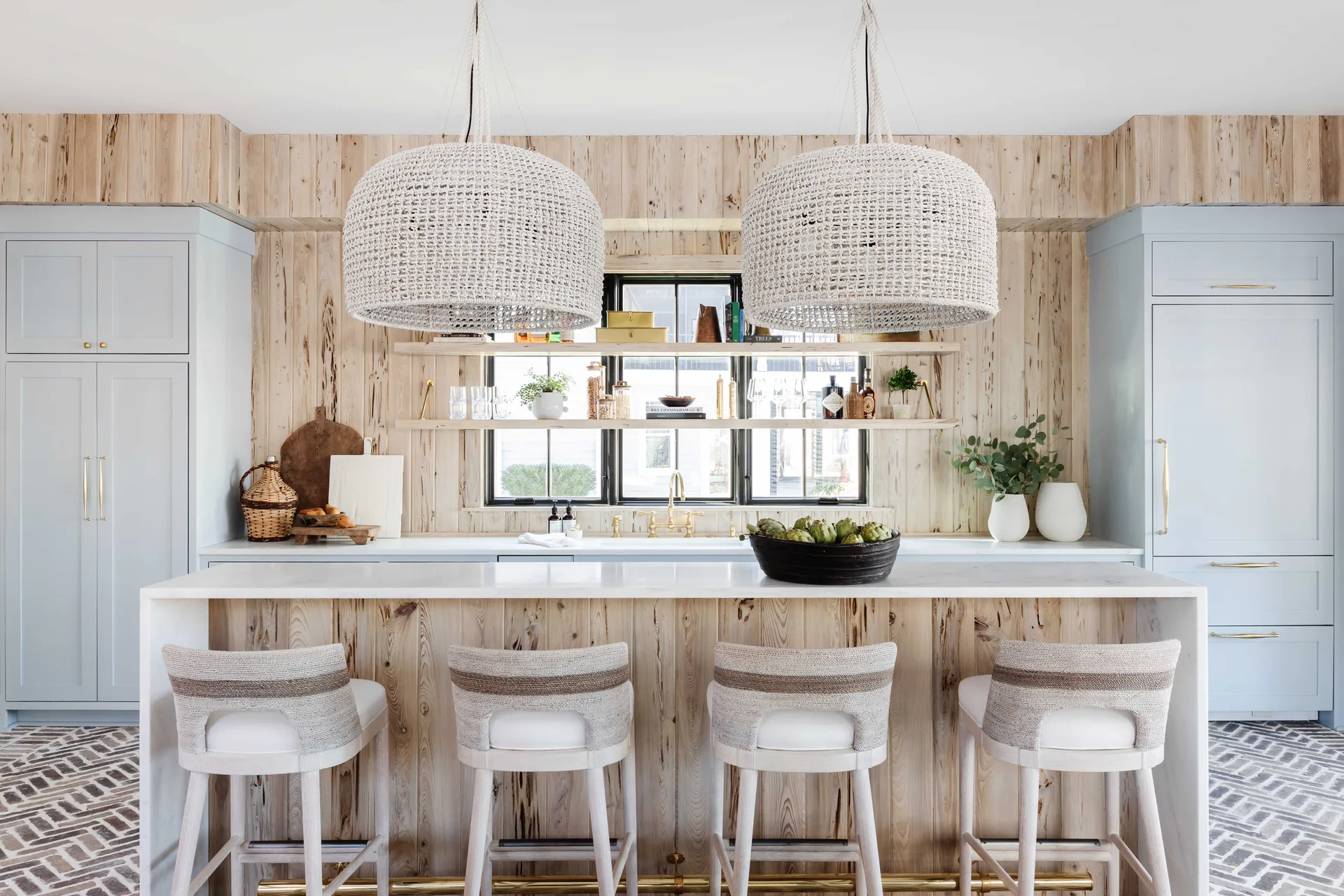 A modern kitchen features a knotty wood-paneled island, pale blue cabinets, and two oversized woven dome pendant lights hanging above the white counter.