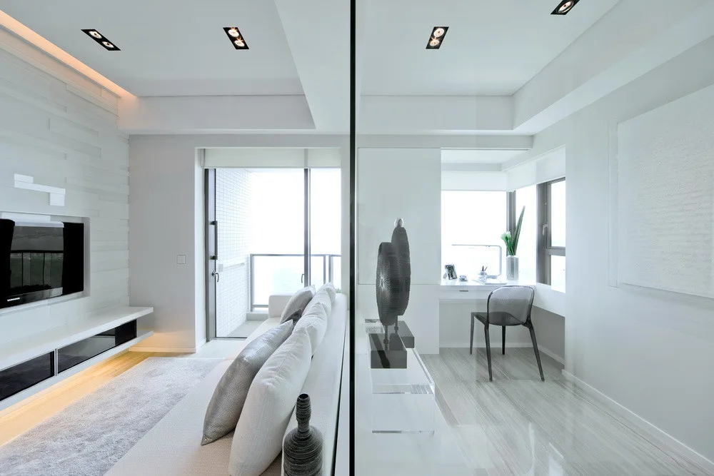A minimalist white living room with a grey sofa and television is separated from a study nook by a clear glass wall.