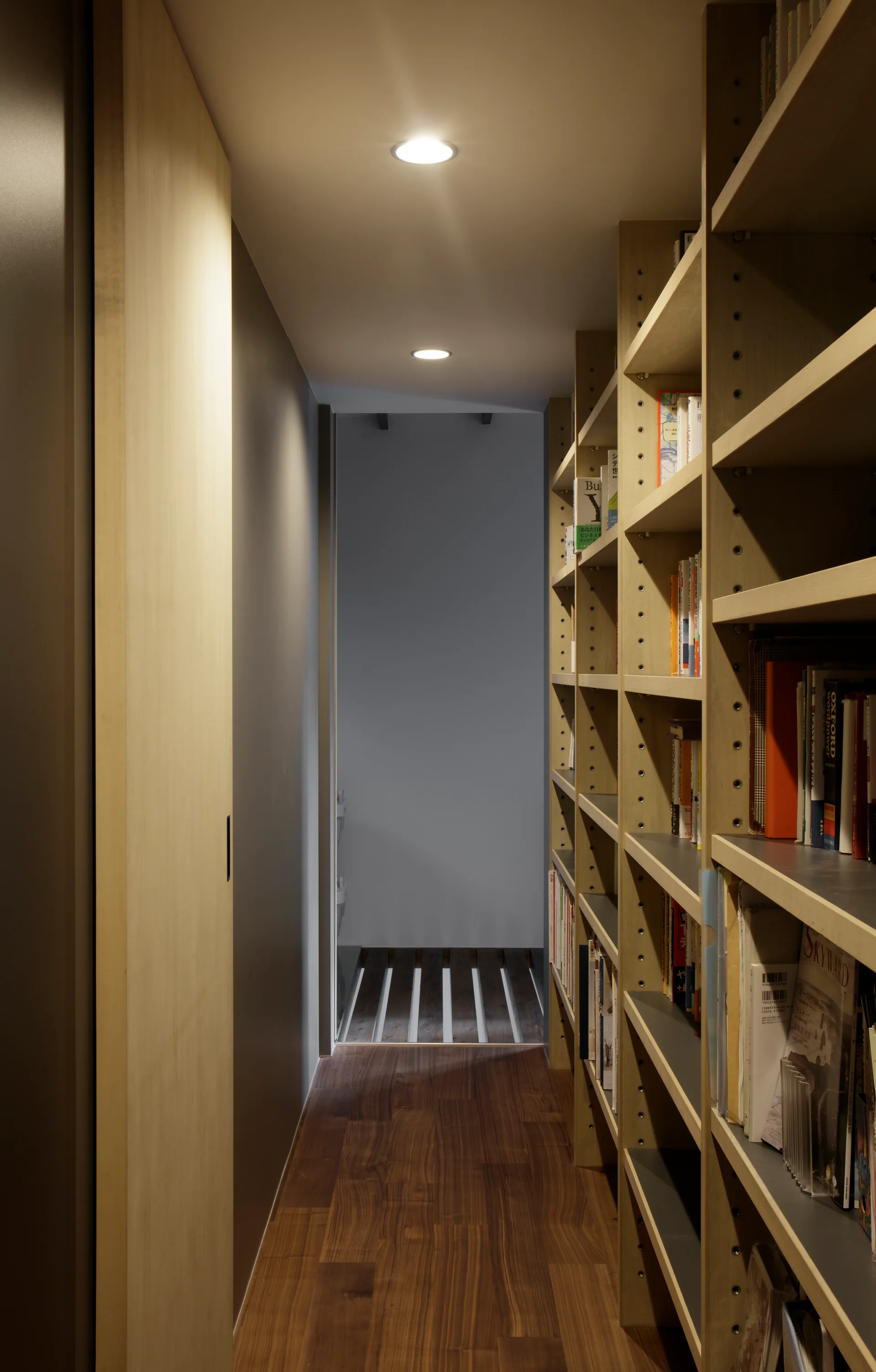 A narrow residential hallway features a tall built-in wooden bookshelf, a sliding wood door, and recessed ceiling lights.