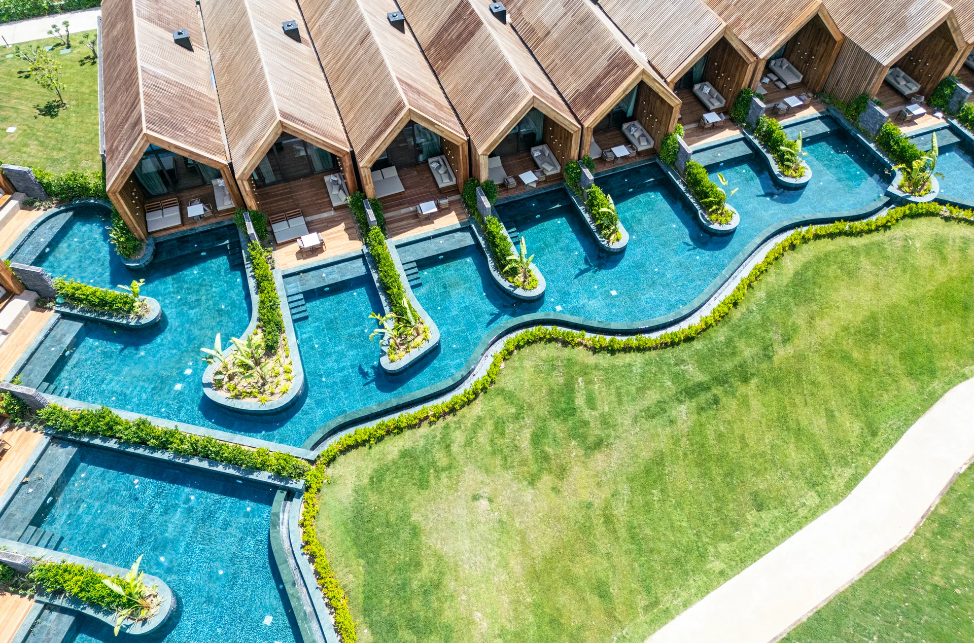Aerial view of modern wood-slatted villas with gabled roofs and private decks lining a curving, interconnected turquoise pool.