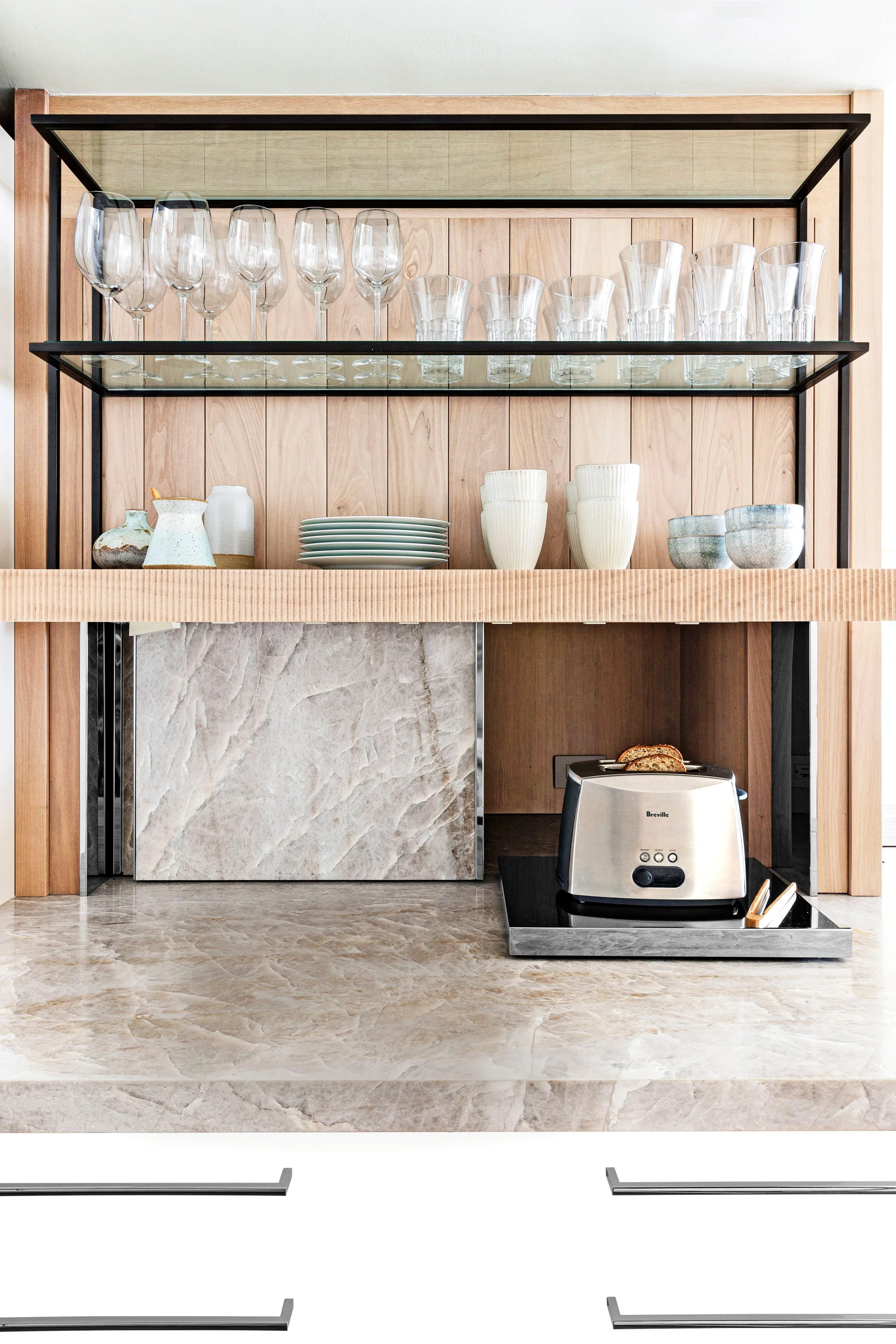 A kitchen niche features black metal-framed glass shelving displaying glassware against a wood-paneled wall above a veined stone countertop.