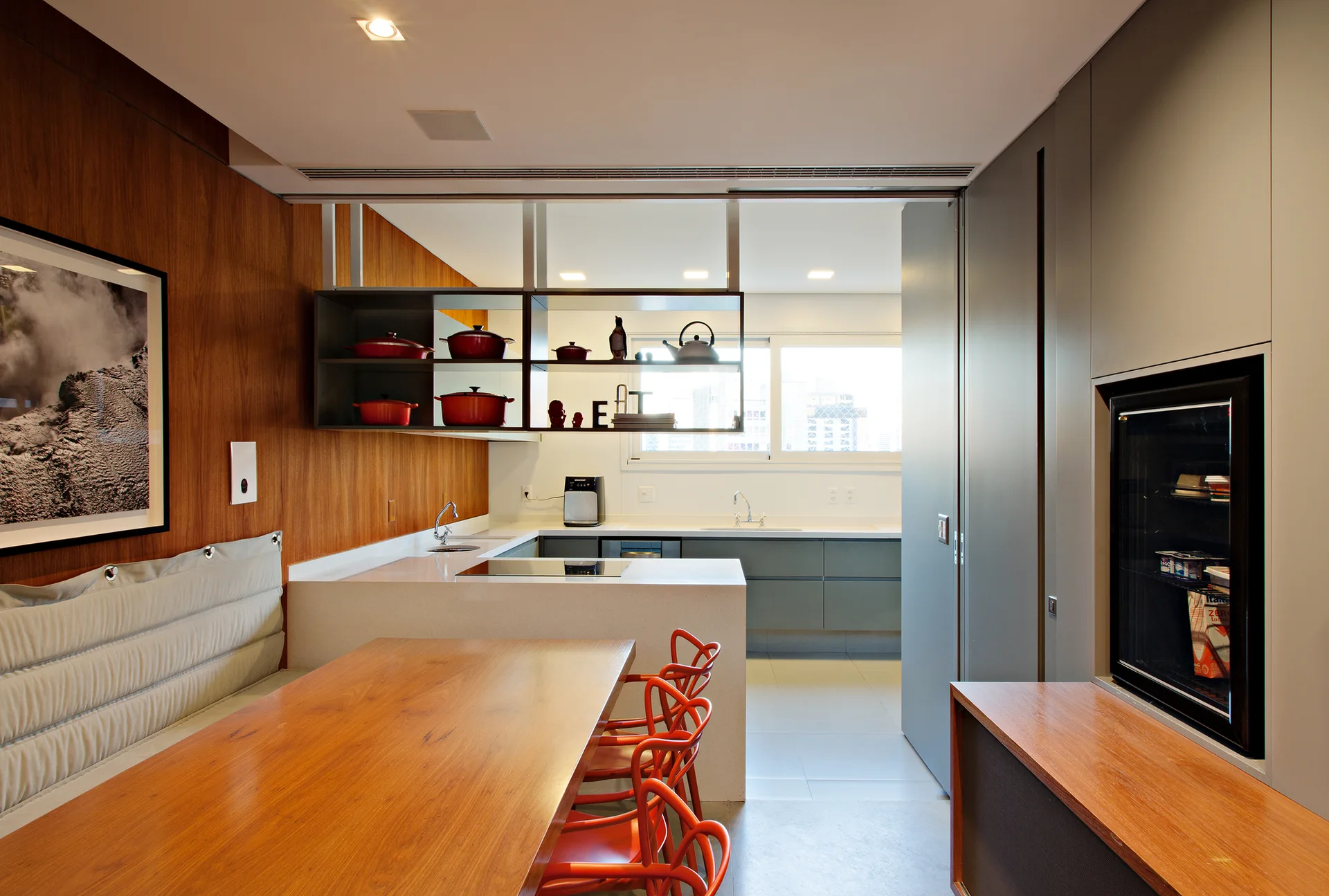 A contemporary kitchen and dining space featuring a wooden dining table, bright red chairs, wood wall paneling, and grey built-in cabinets.