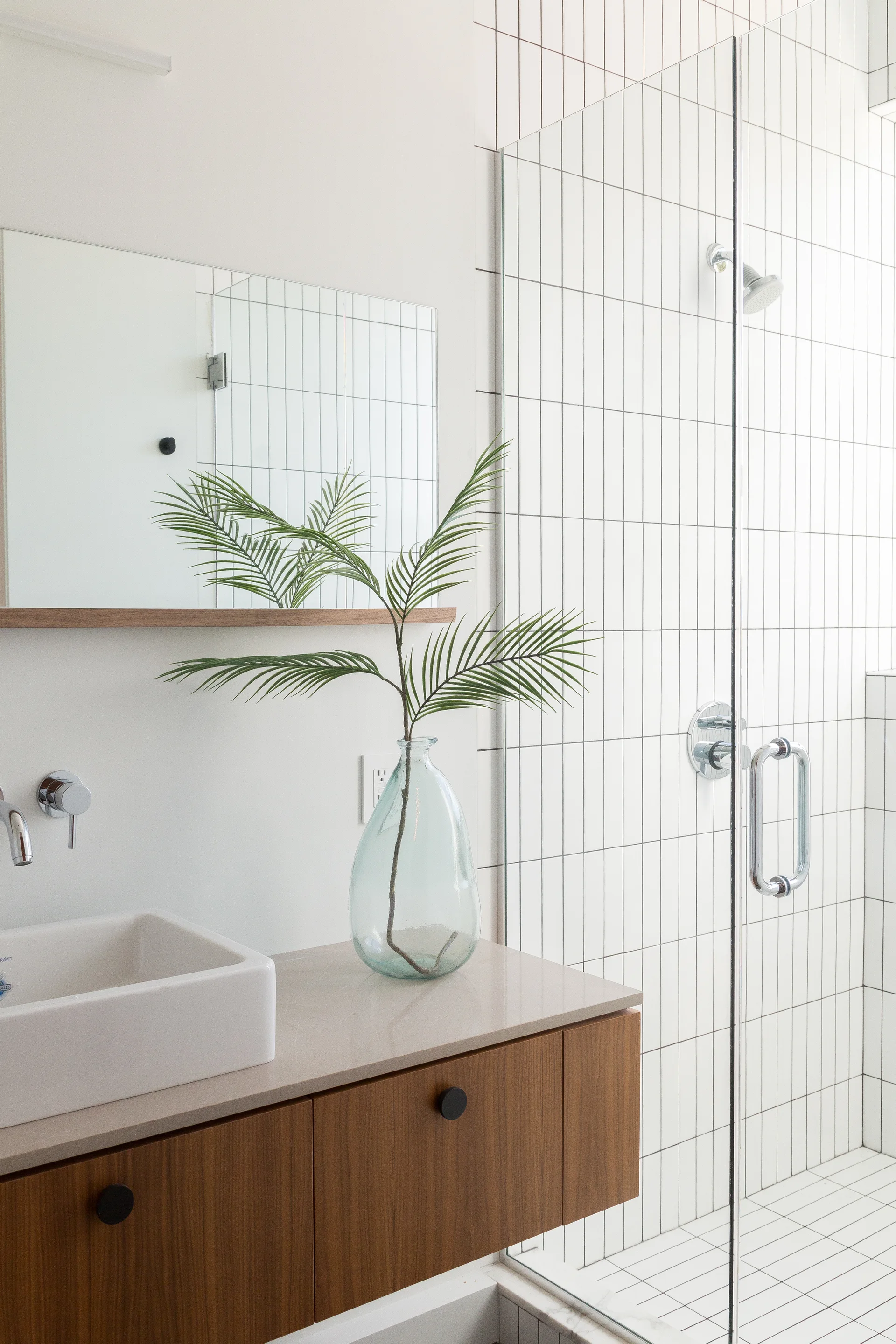 A modern bathroom with a wood floating vanity, a white vessel sink, and a glass shower enclosure with vertically stacked white subway tiles.