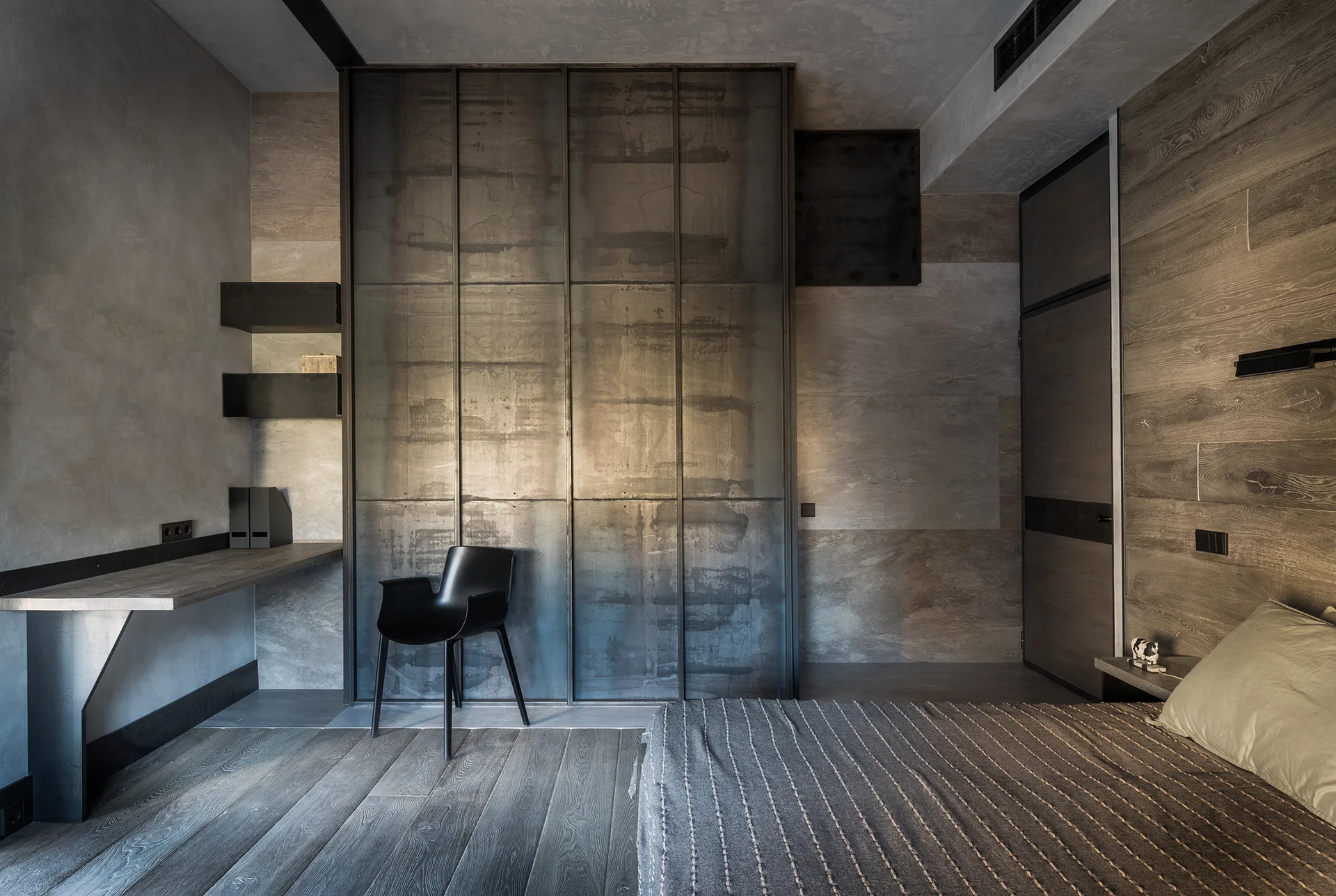 An industrial bedroom with an oxidized metal wardrobe, a floating wooden desk, a black armchair, and a bed with textured linens.