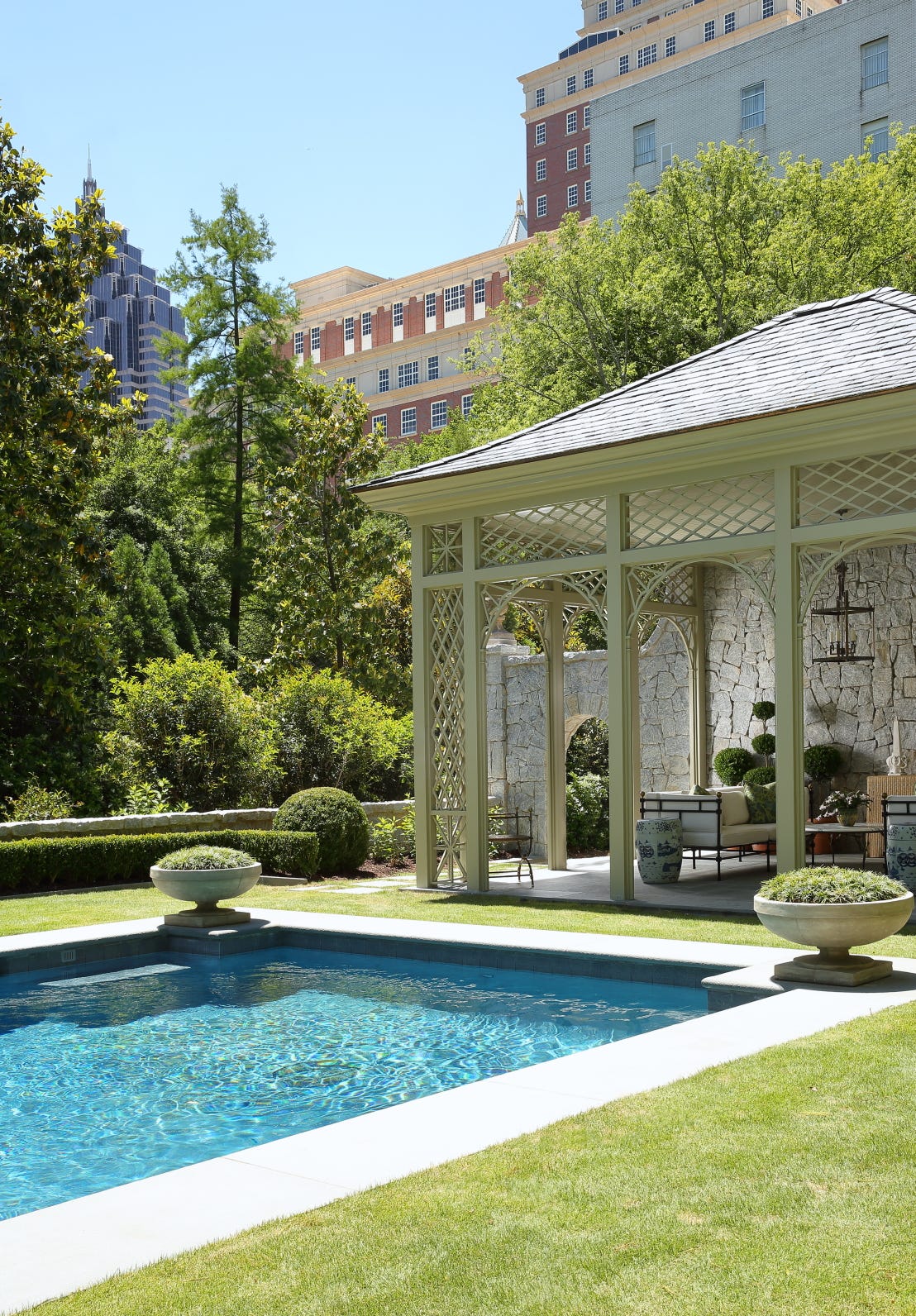 A formal swimming pool sits beside a pale green latticed pavilion featuring an outdoor sofa, stone walls, and blue-and-white garden stools.