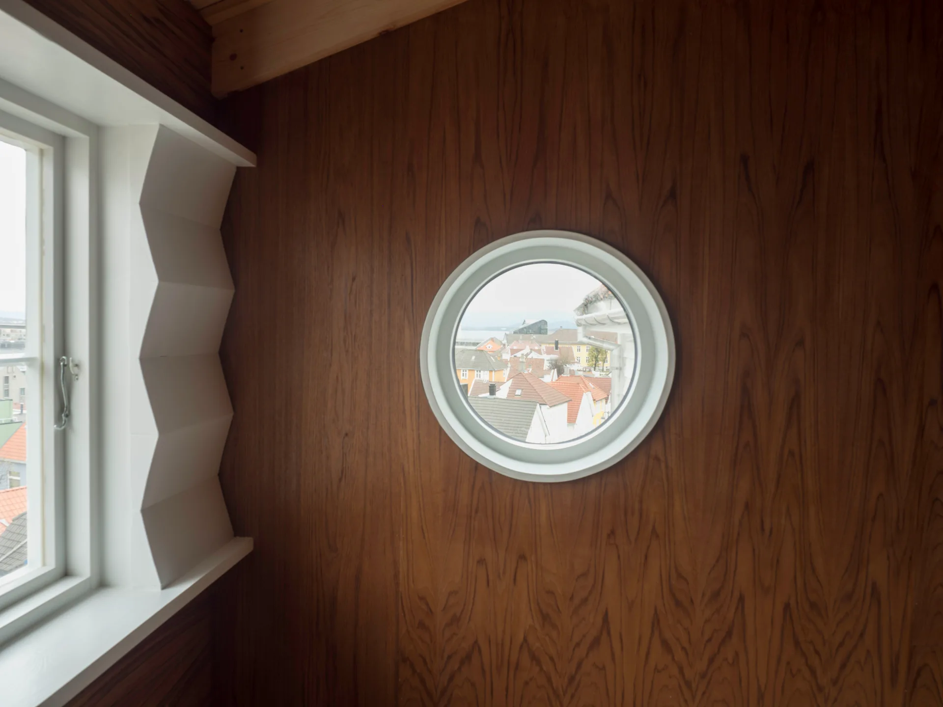 A round porthole window and a geometric white window frame are set into a warm wood-paneled wall overlooking rooftops.