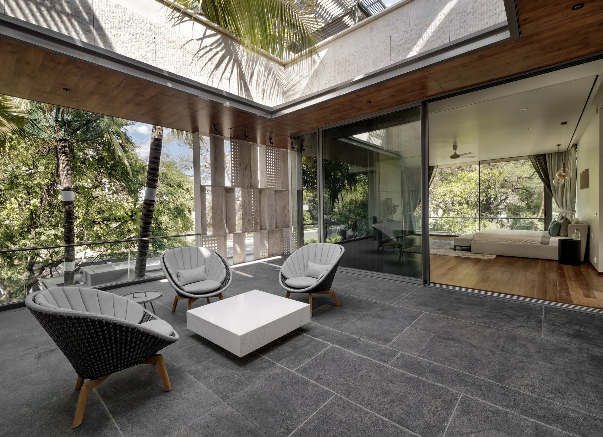 A modern covered patio with grey stone flooring features three grey lounge chairs, a white coffee table, and large sliding glass doors leading to a bedroom.