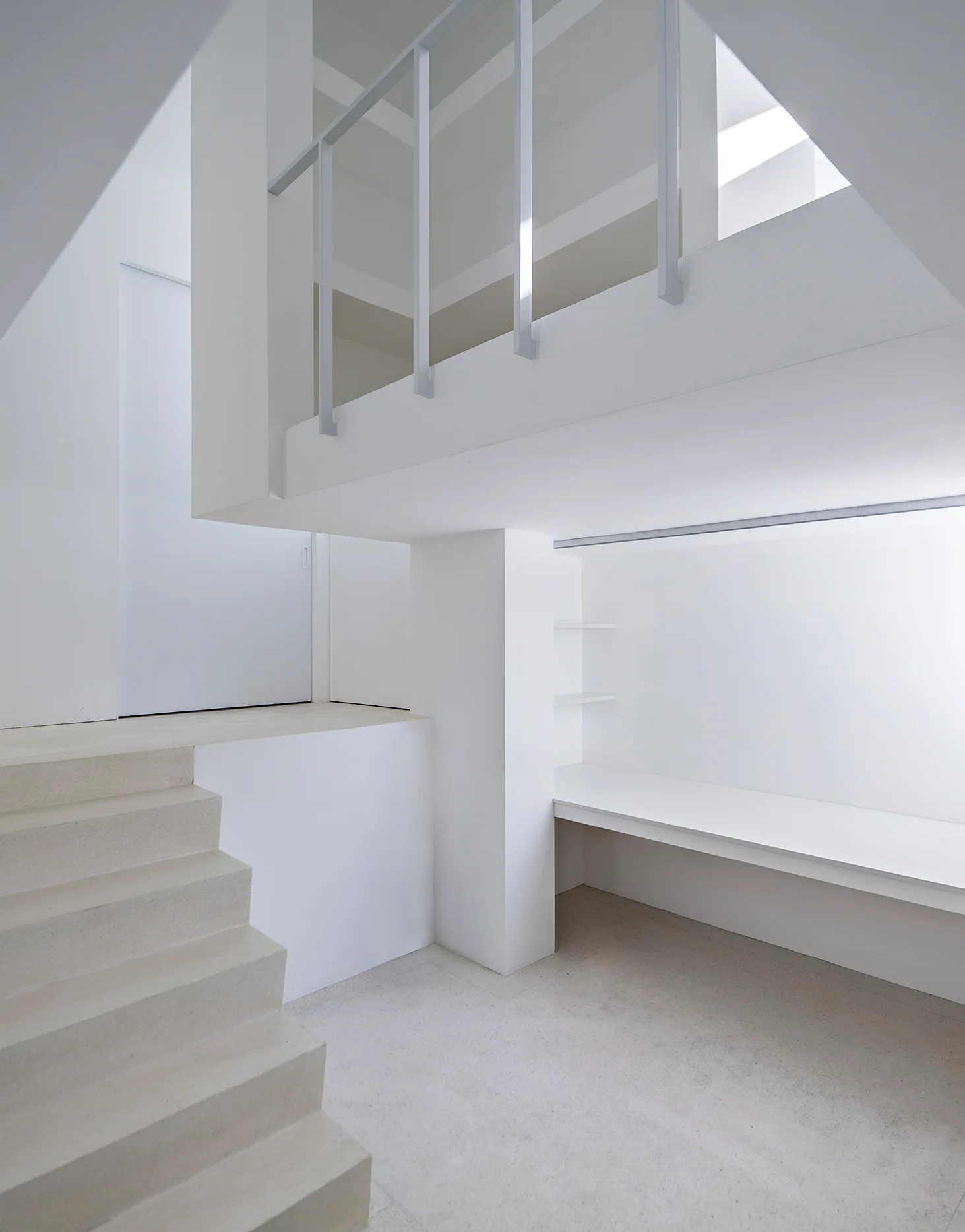 An all-white minimalist interior featuring a plaster staircase, a built-in desk nestled under the upper level, and a simple metal railing.