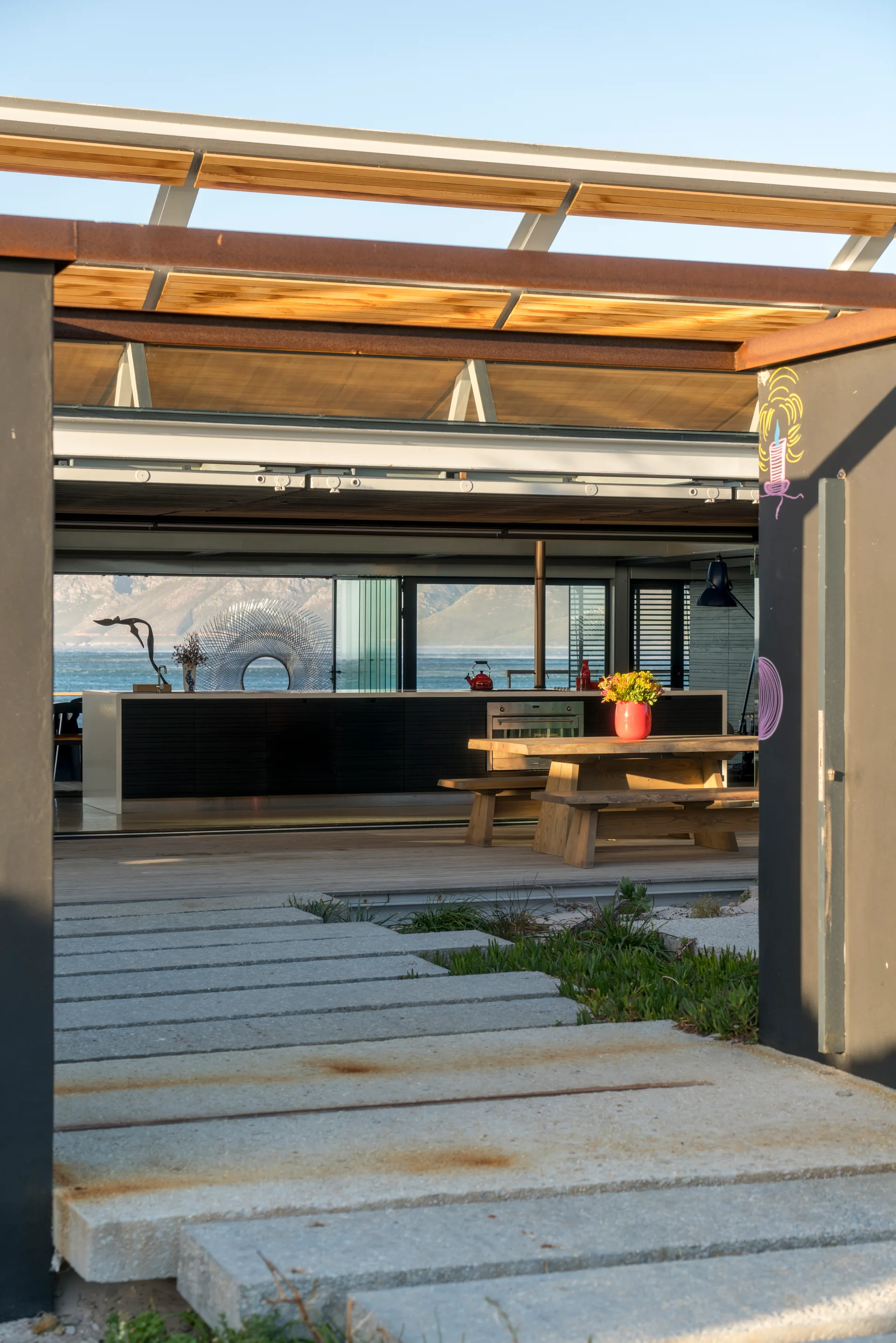 Large concrete stepping stones lead toward an open-concept wooden patio and indoor kitchen overlooking a coastal ocean view.
