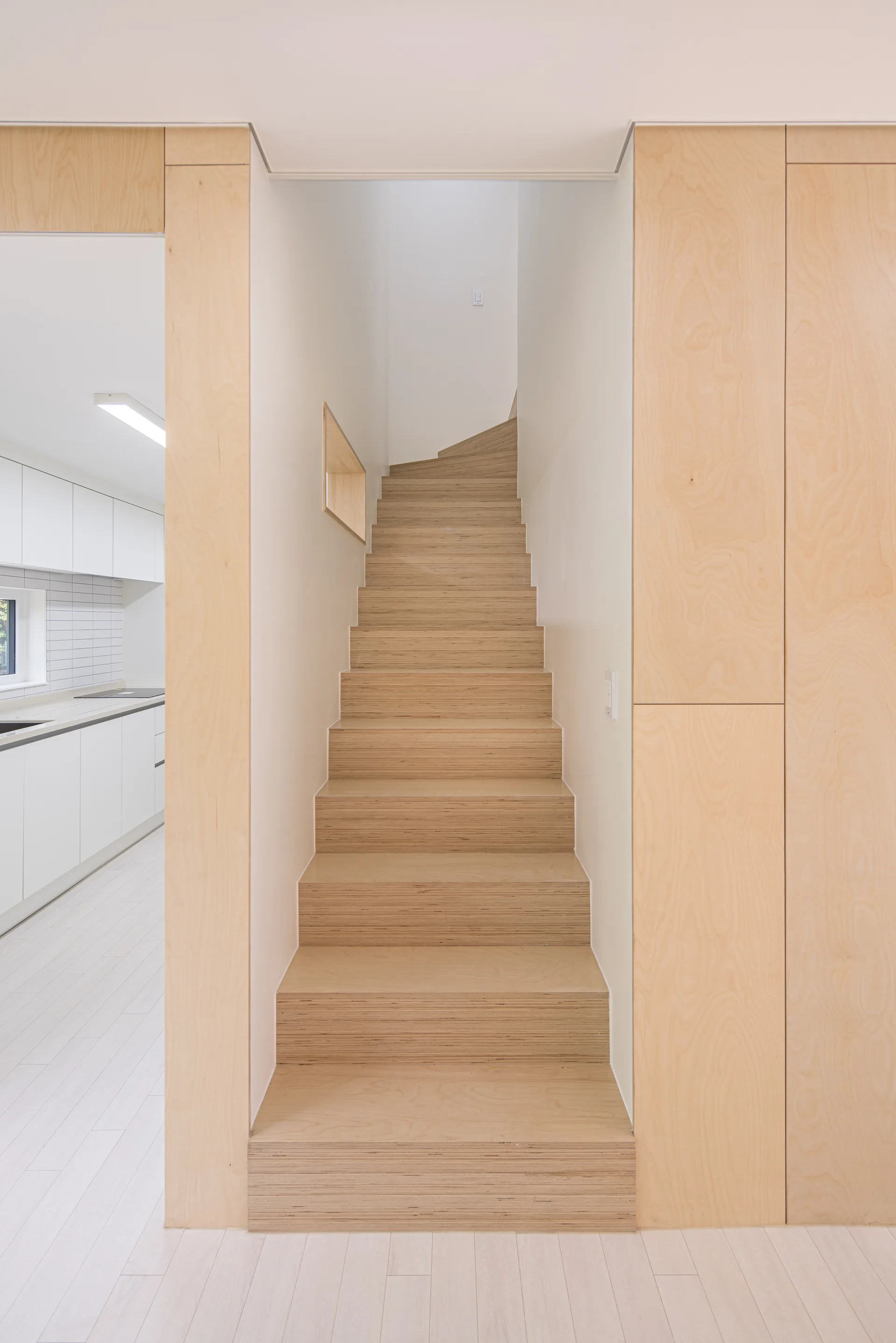 A minimalist light wood staircase is flanked by seamless plywood wall panels next to a bright white kitchen.