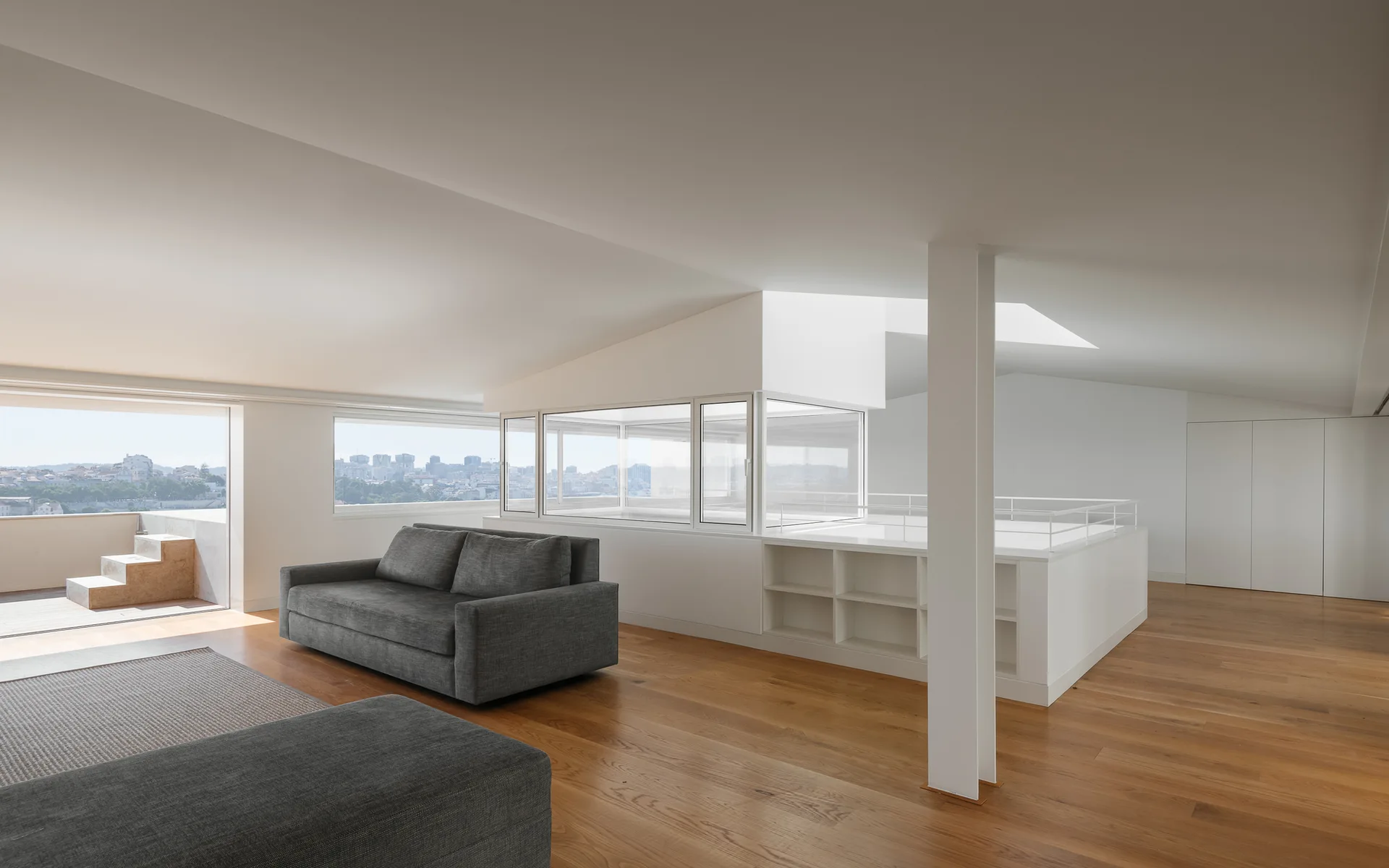 A minimalist loft living room features a grey fabric sofa, light wood flooring, and a central white built-in shelving unit beneath a glass enclosure.