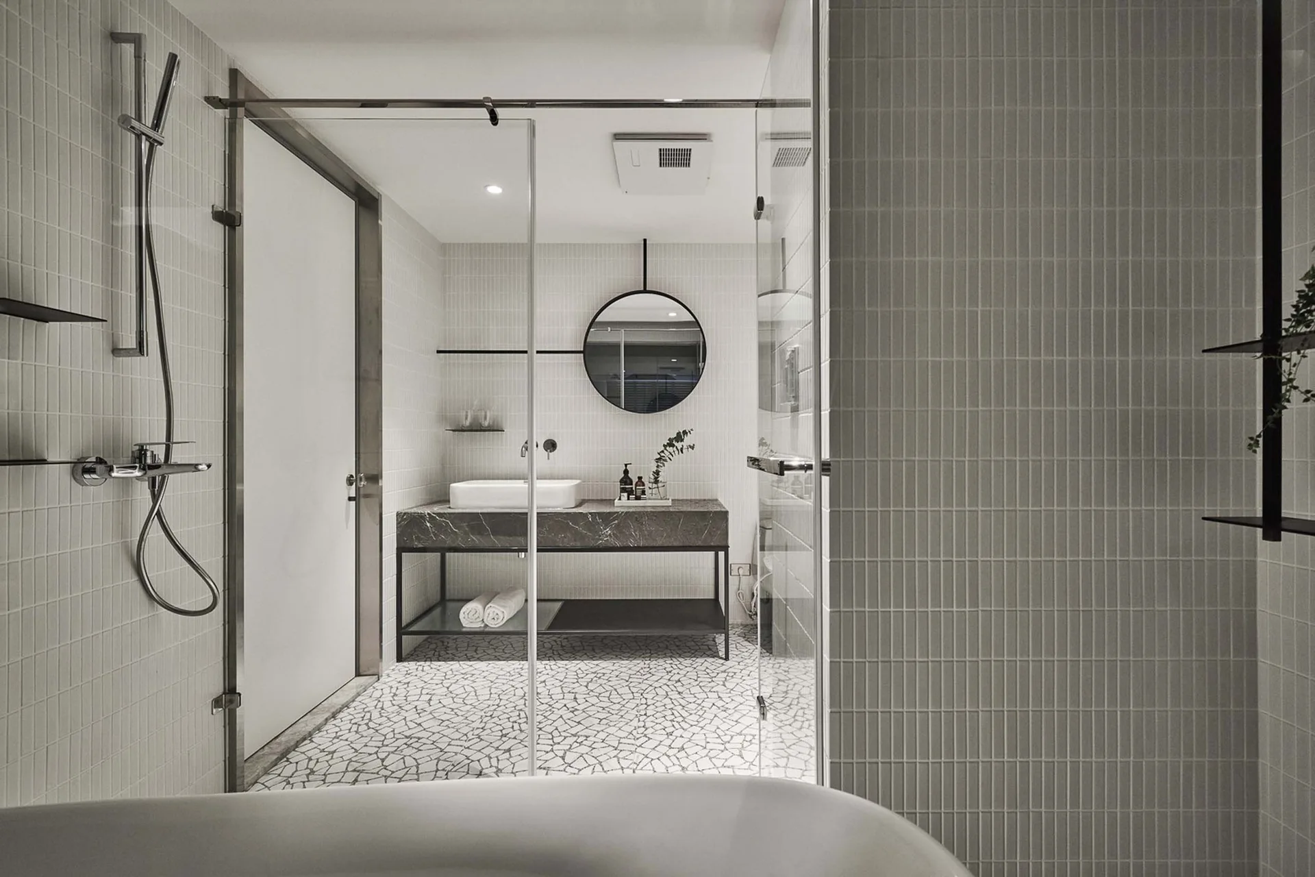 A minimalist bathroom featuring a dark stone vanity with a circular mirror, vertical white wall tiles, and a black and white cracked mosaic floor.
