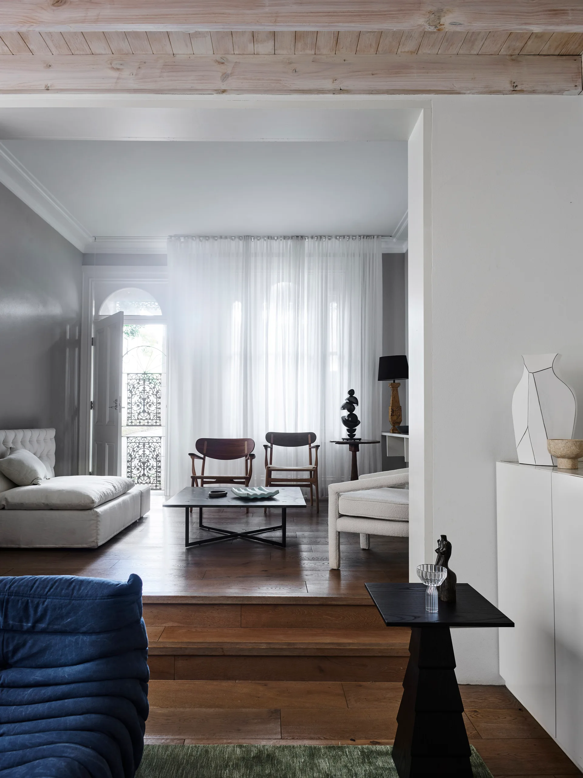 A split-level living room features modern wooden chairs, a low coffee table, and a white sofa set against sheer drapes.