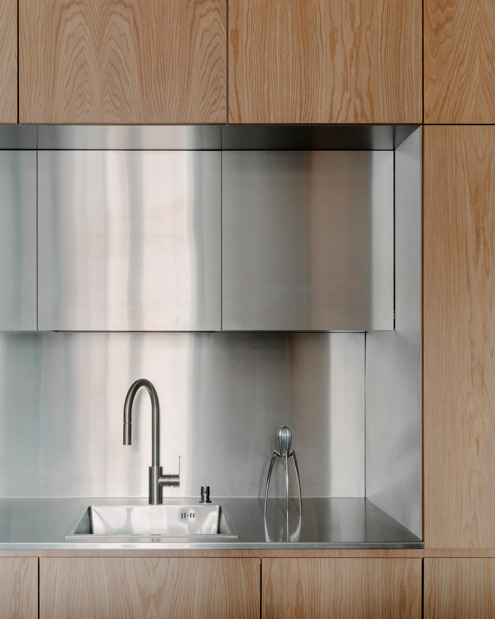 A minimalist kitchen sink with a gooseneck faucet is integrated into a stainless steel countertop and backsplash framed by light wood cabinets.