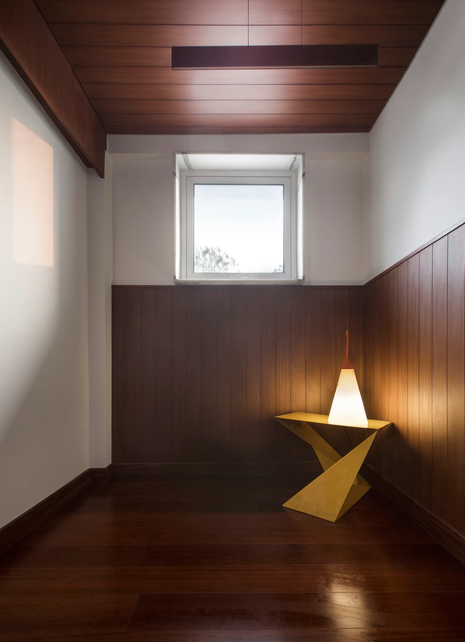 A corner nook features dark wood-paneled walls and flooring, a central window, and an illuminated cone lamp sitting on a bright yellow geometric table.