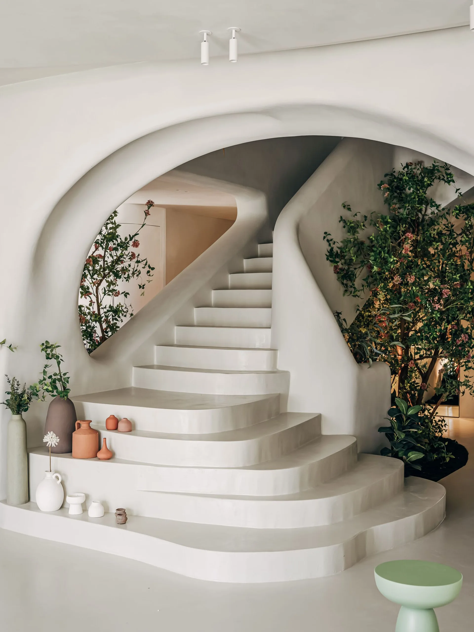 A curved white plaster staircase with a built-in archway, decorated with terracotta ceramic vases and indoor green foliage.