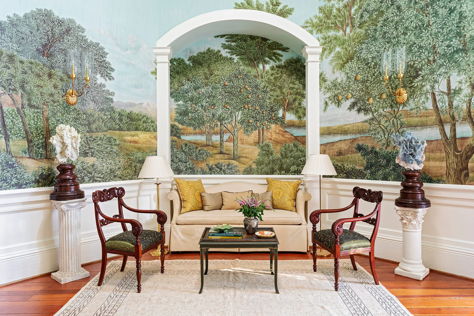 A traditional sitting area with a cream sofa set within a white archway against a scenic landscape wall mural flanked by two dark wood armchairs.