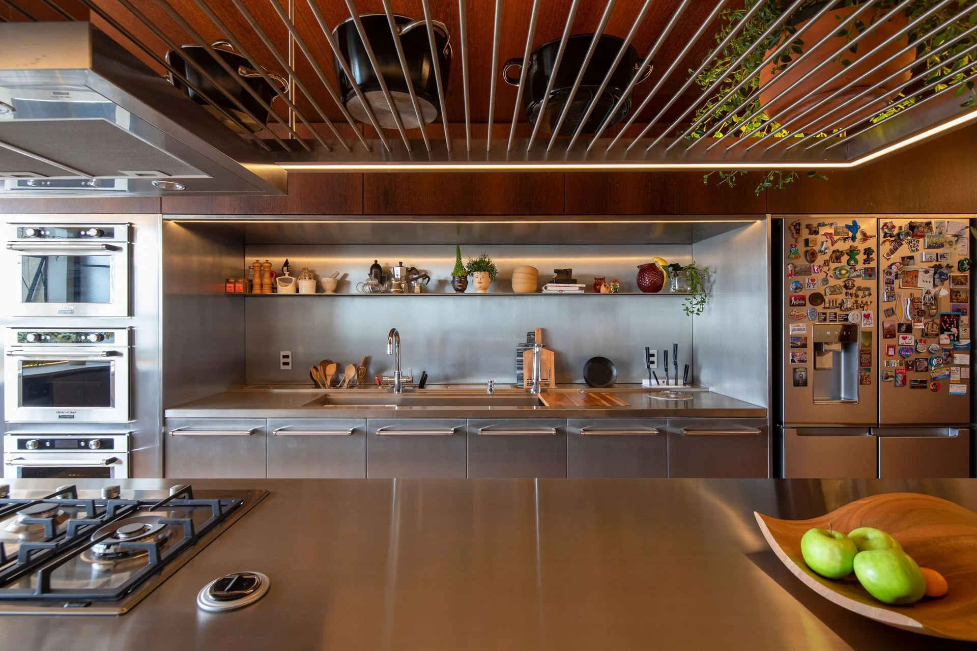 A modern kitchen featuring stainless steel countertops, a metallic backsplash, built-in appliances, and a slatted wood ceiling with a suspended pot rack.
