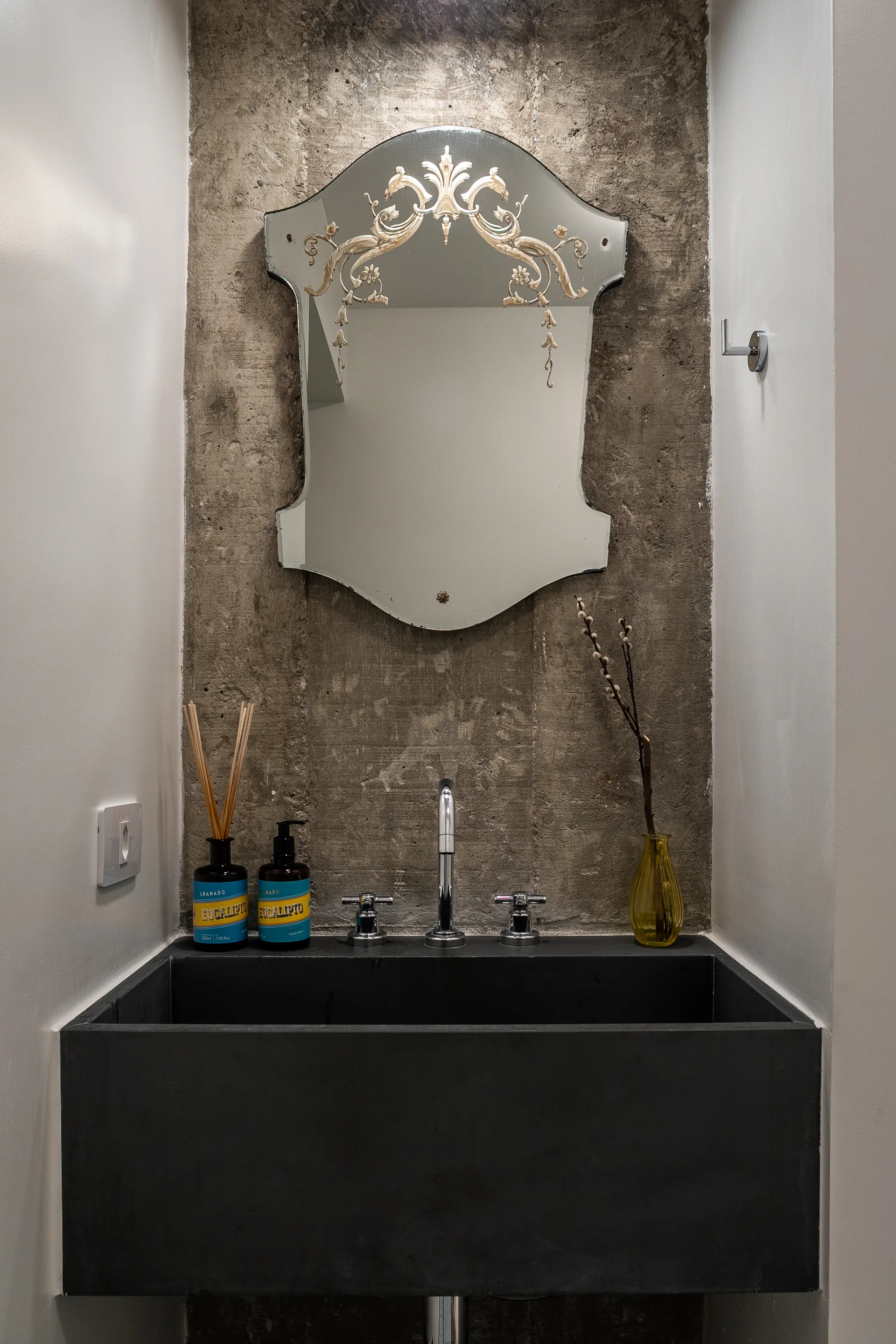 An ornate etched glass mirror hangs on a raw concrete wall above a wide matte black trough sink in a minimalist powder room.