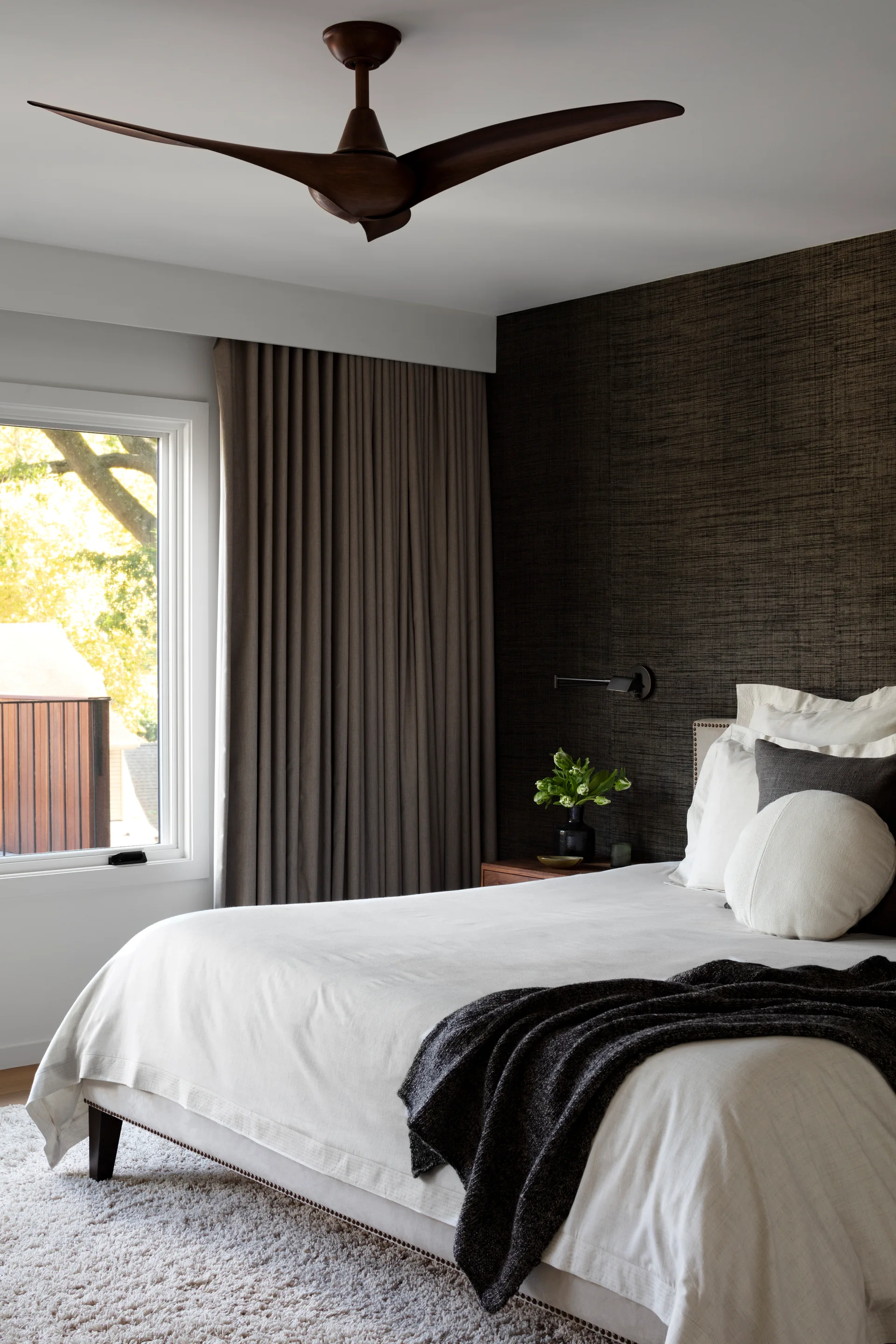 A modern bedroom features a white bed set against a dark textured accent wall next to a window with long grey curtains and a wooden ceiling fan.