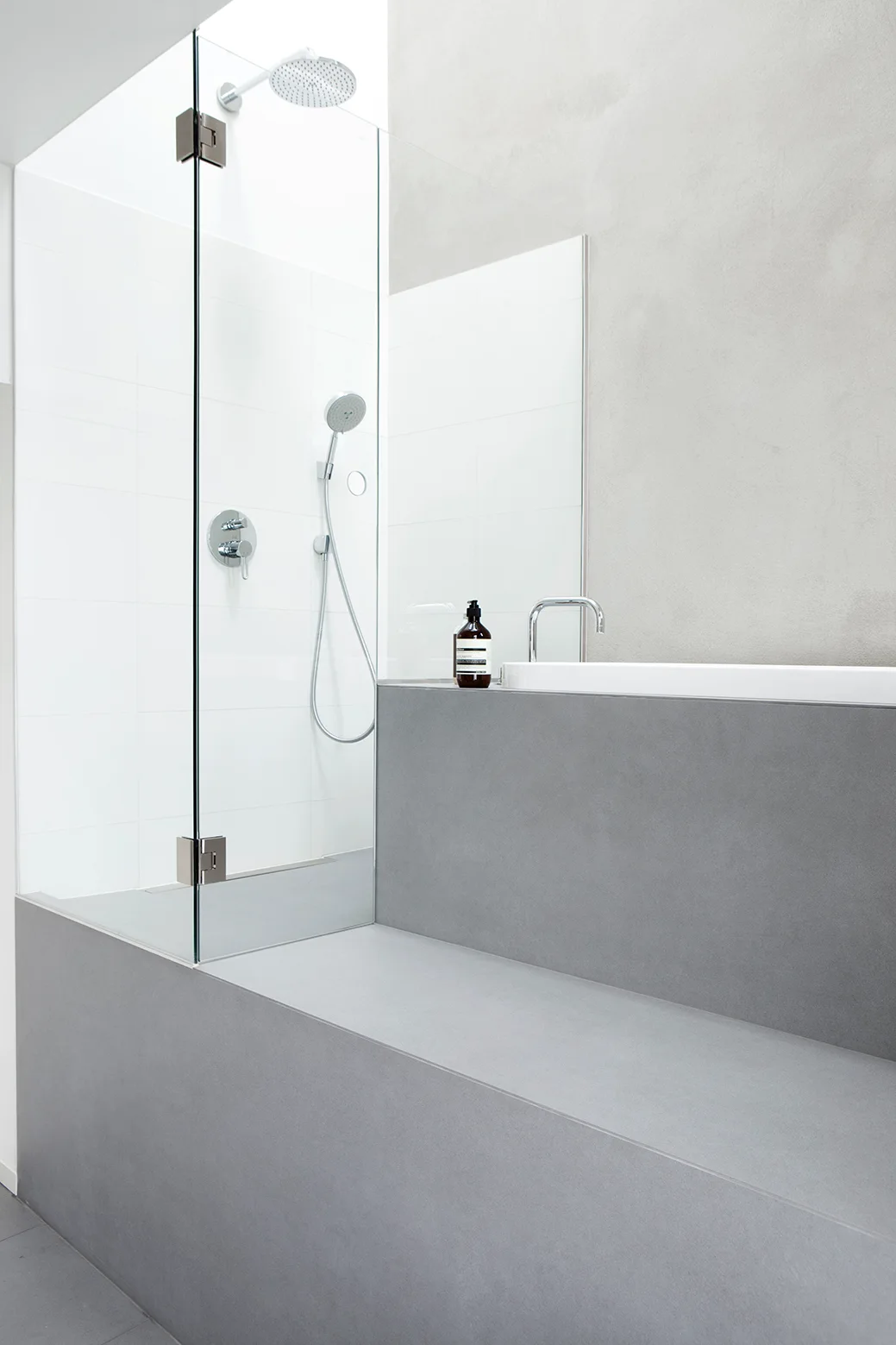 A minimalist bathroom features a clear glass shower partition next to a built-in bathtub surrounded by smooth matte grey stone panels.