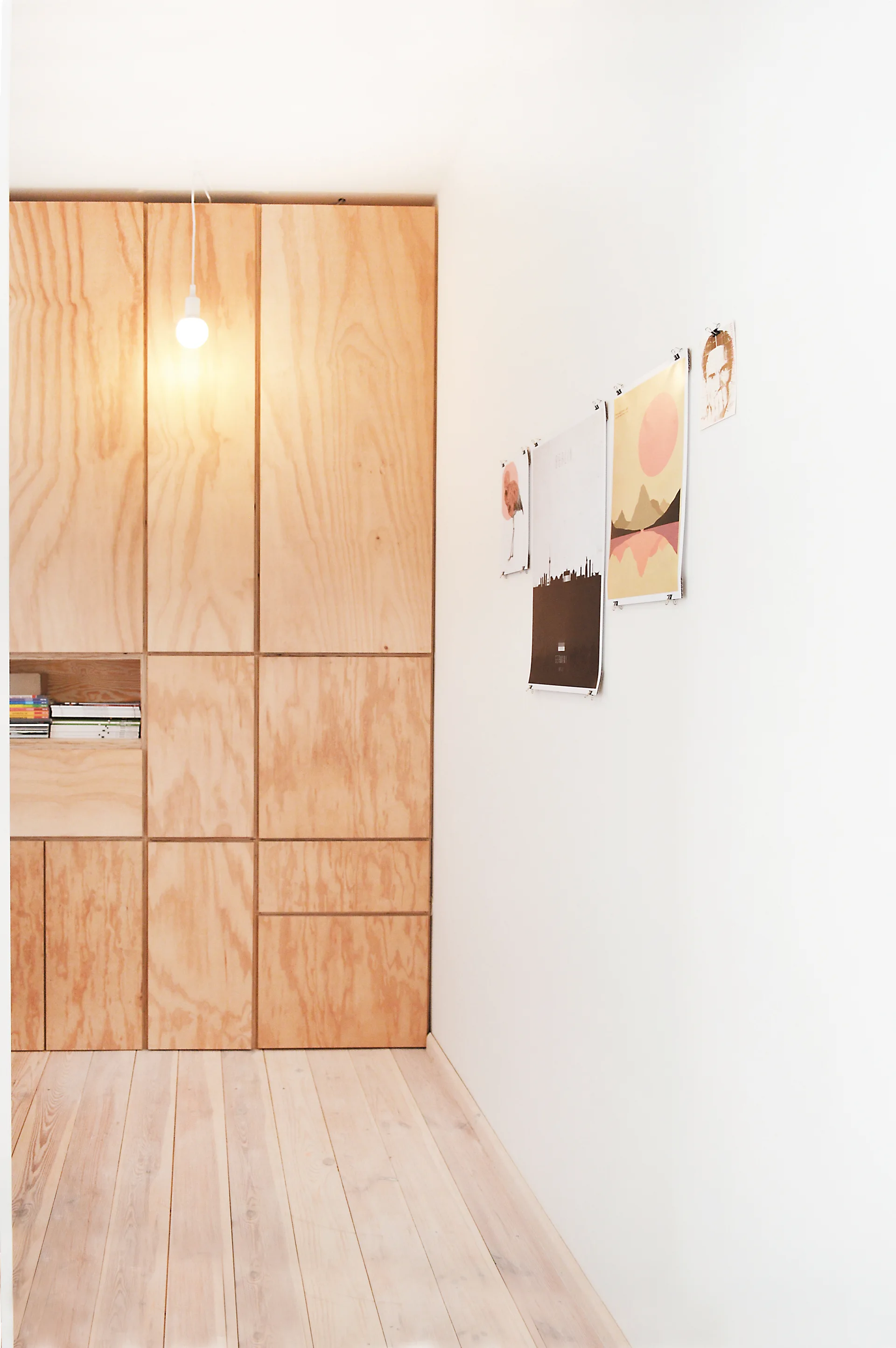 A minimalist room corner features custom floor-to-ceiling plywood cabinets, light wood flooring, a single pendant light, and prints clipped to a white wall.