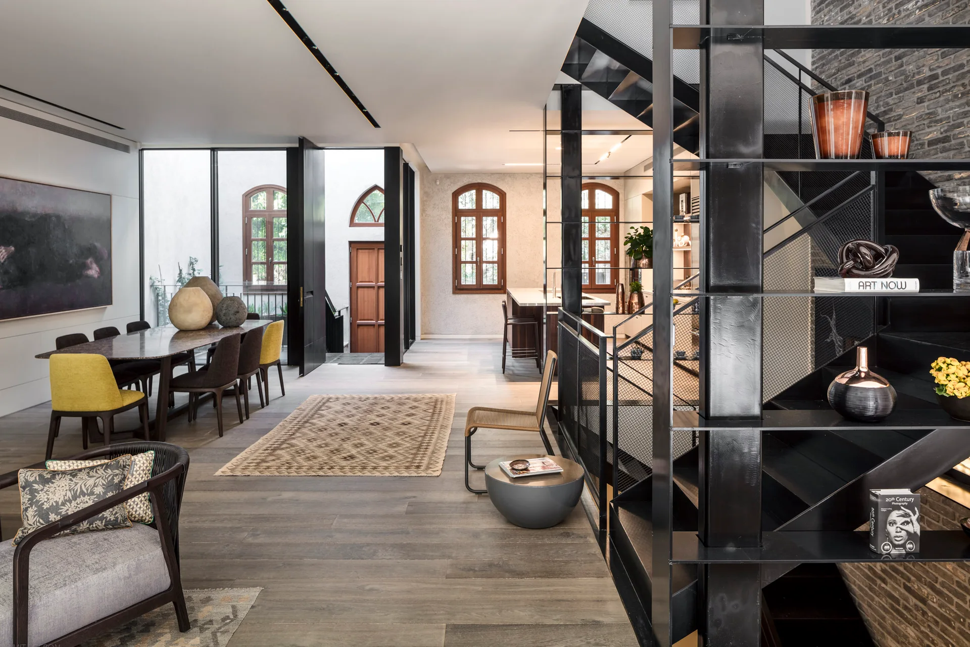 A modern open interior featuring a prominent black metal staircase, a wood dining table with yellow chairs, and arched wooden windows.