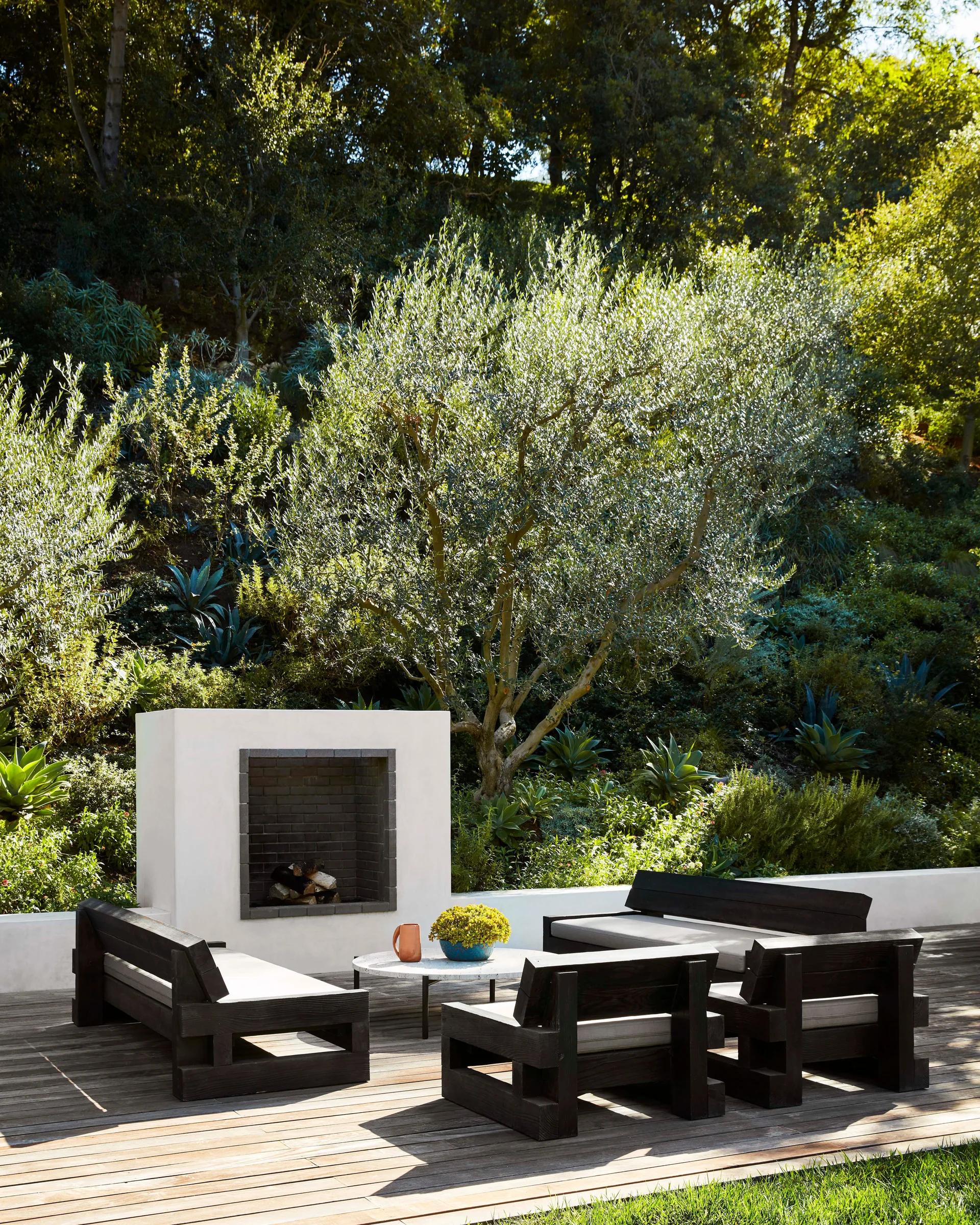 A modern outdoor patio features a white stucco fireplace and black wood lounge furniture on a wooden deck with a lush green garden background.