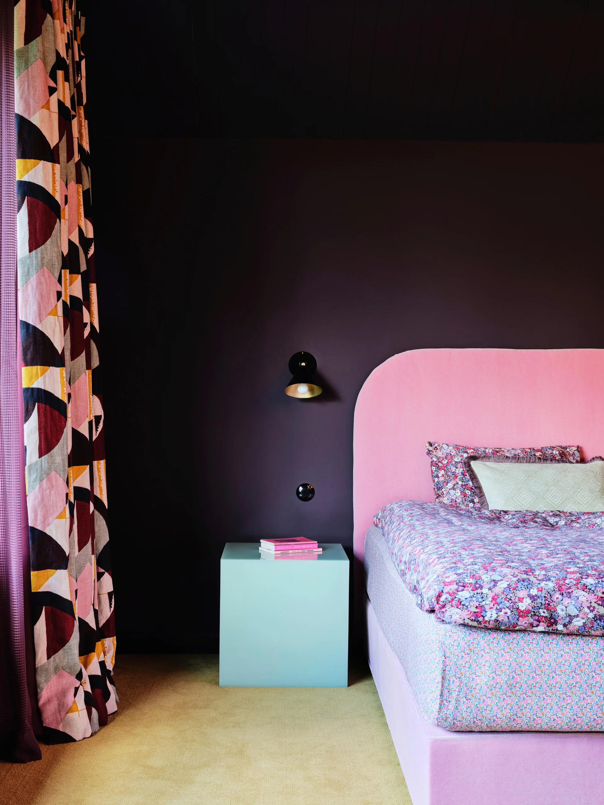 A pink upholstered headboard and floral bedding sit against a dark brown wall next to a light blue cube nightstand.
