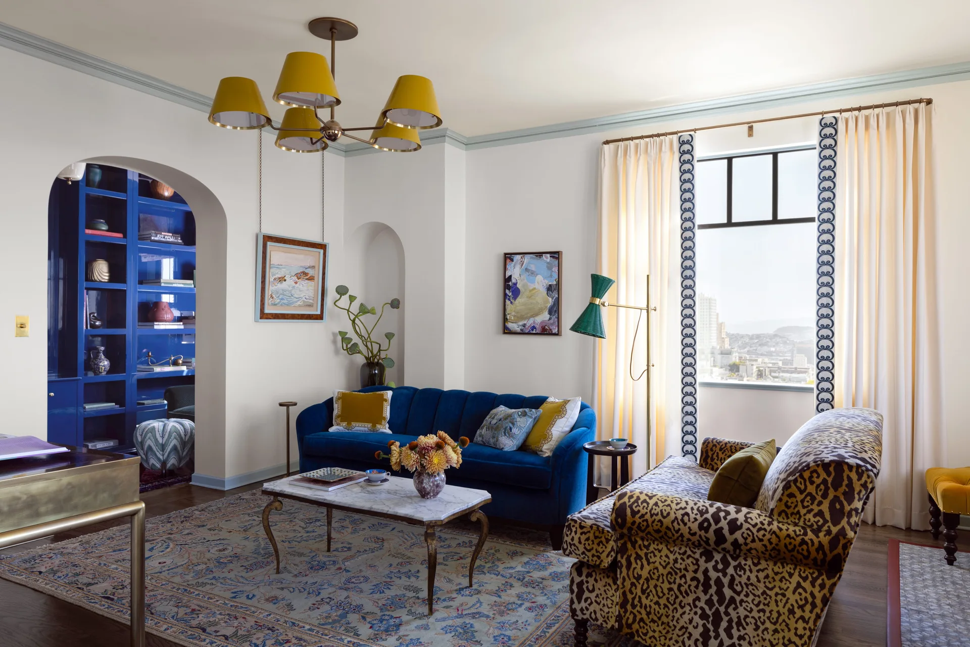 An eclectic living room features a blue velvet sofa, a leopard-print armchair, a marble coffee table, and blue built-in shelving framed by an archway.