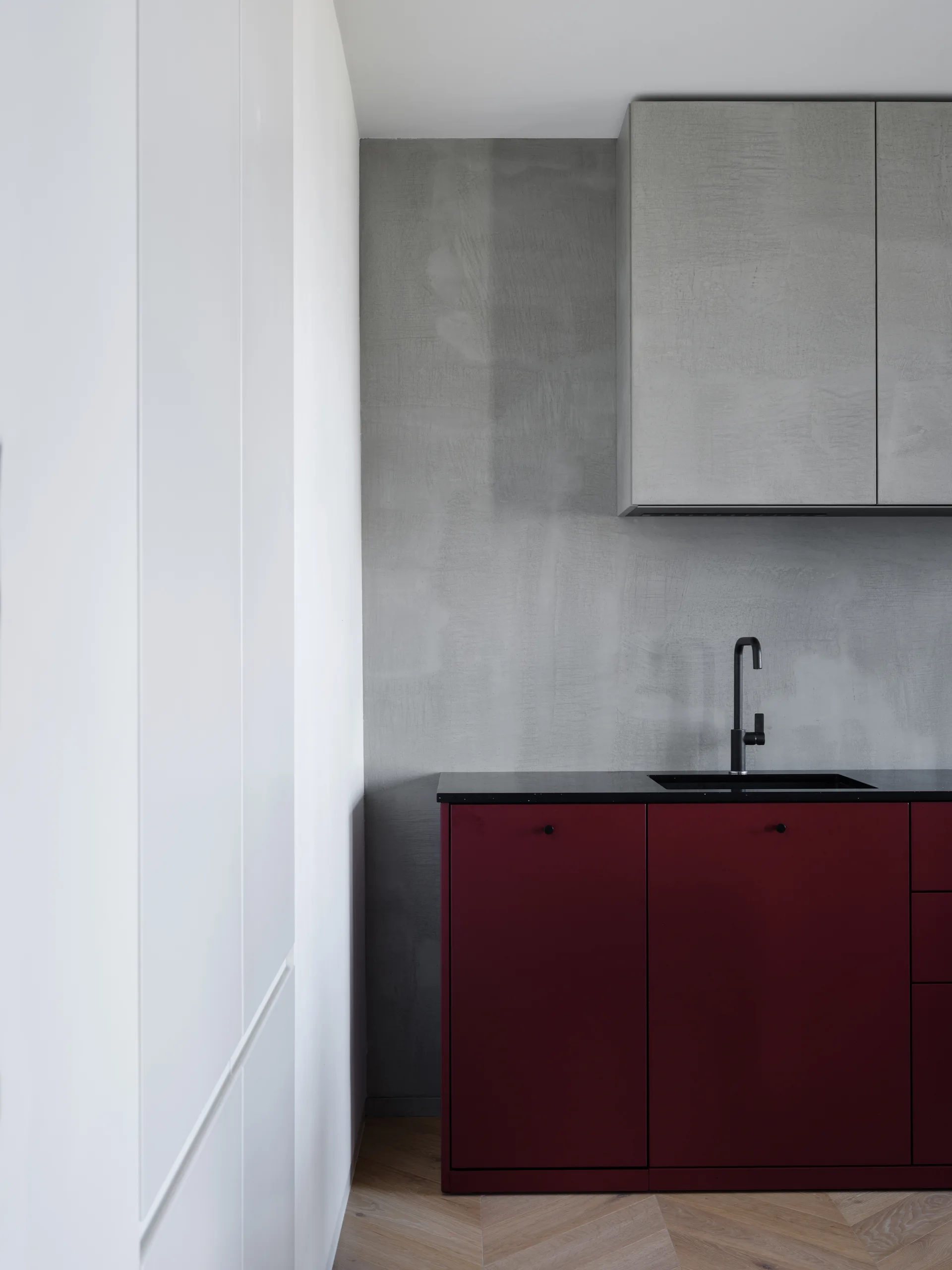 Modern kitchen detail showing deep red lower cabinets, a black sink and faucet, a textured gray plaster backsplash, and light wood herringbone flooring.