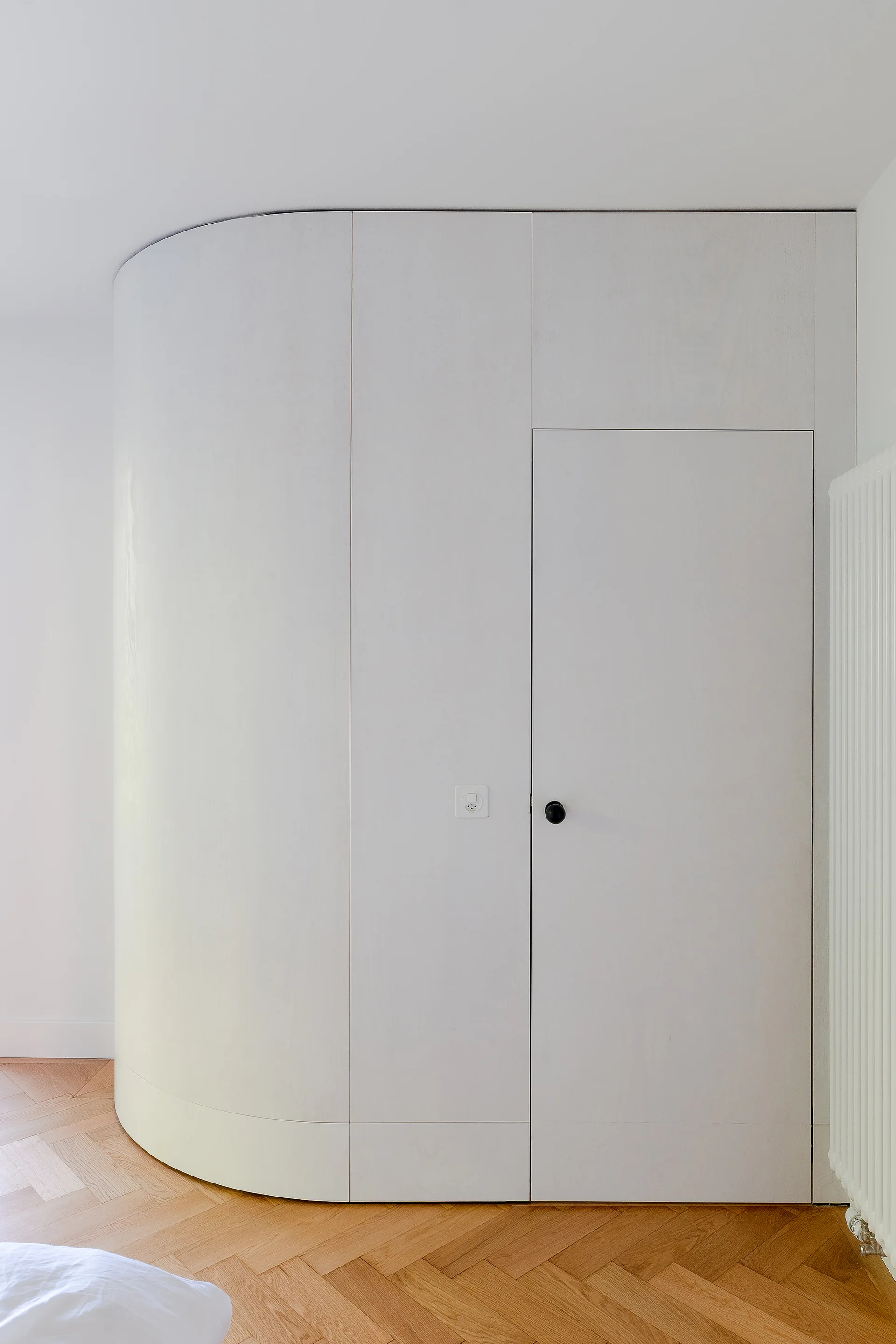 A minimalist white curved wall unit with an integrated door stands on a light oak herringbone parquet floor next to a white radiator.