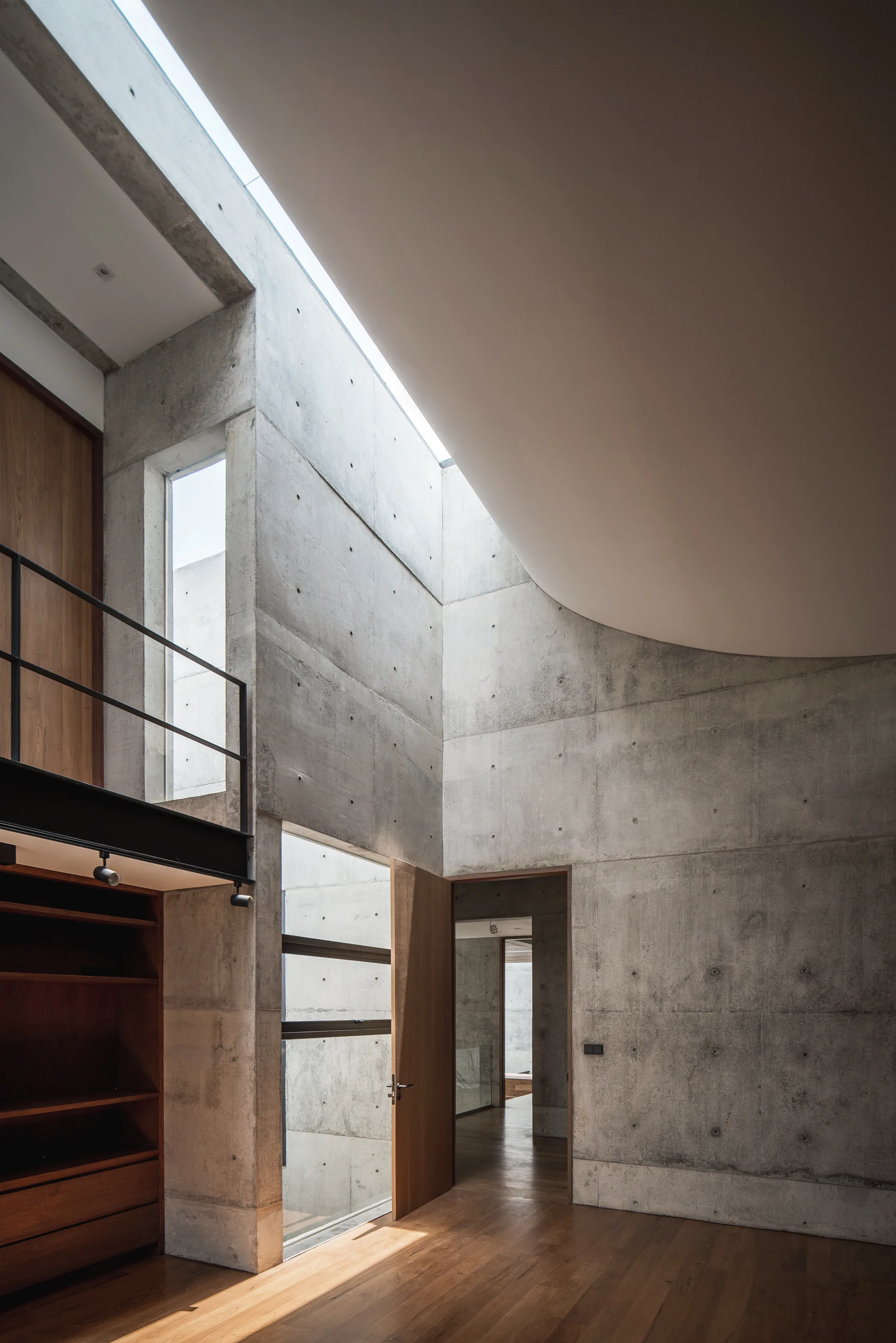 A modern hallway features exposed concrete walls, warm wood flooring, a black metal mezzanine railing, and light from a narrow skylight.