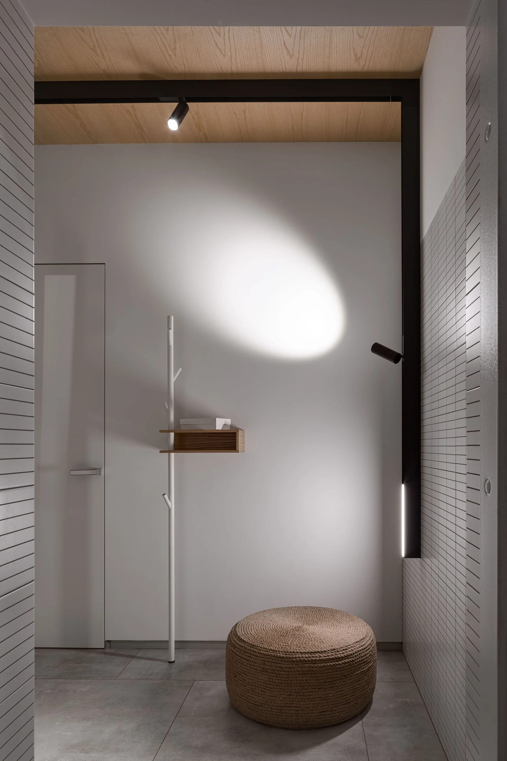 A minimalist entryway features a slim white coat rack with an attached wood shelf, a round woven pouf, and a wood plank ceiling.