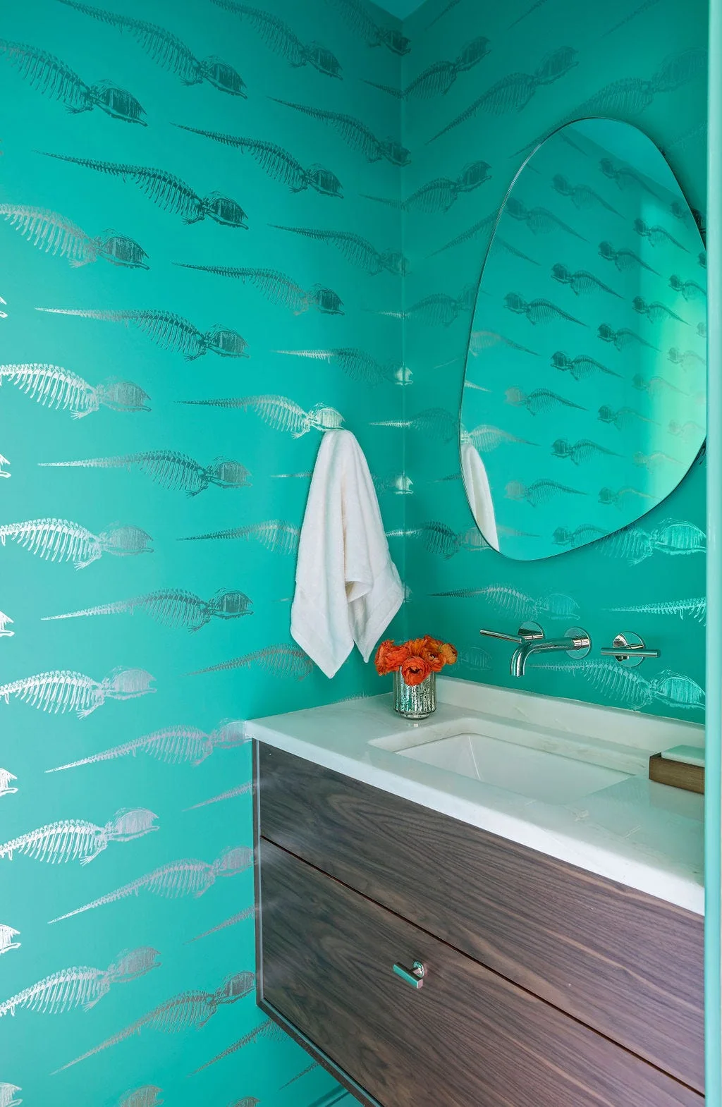 A teal powder room with metallic skeleton wallpaper, a floating wood vanity with integrated sink, and an organic-shaped mirror.