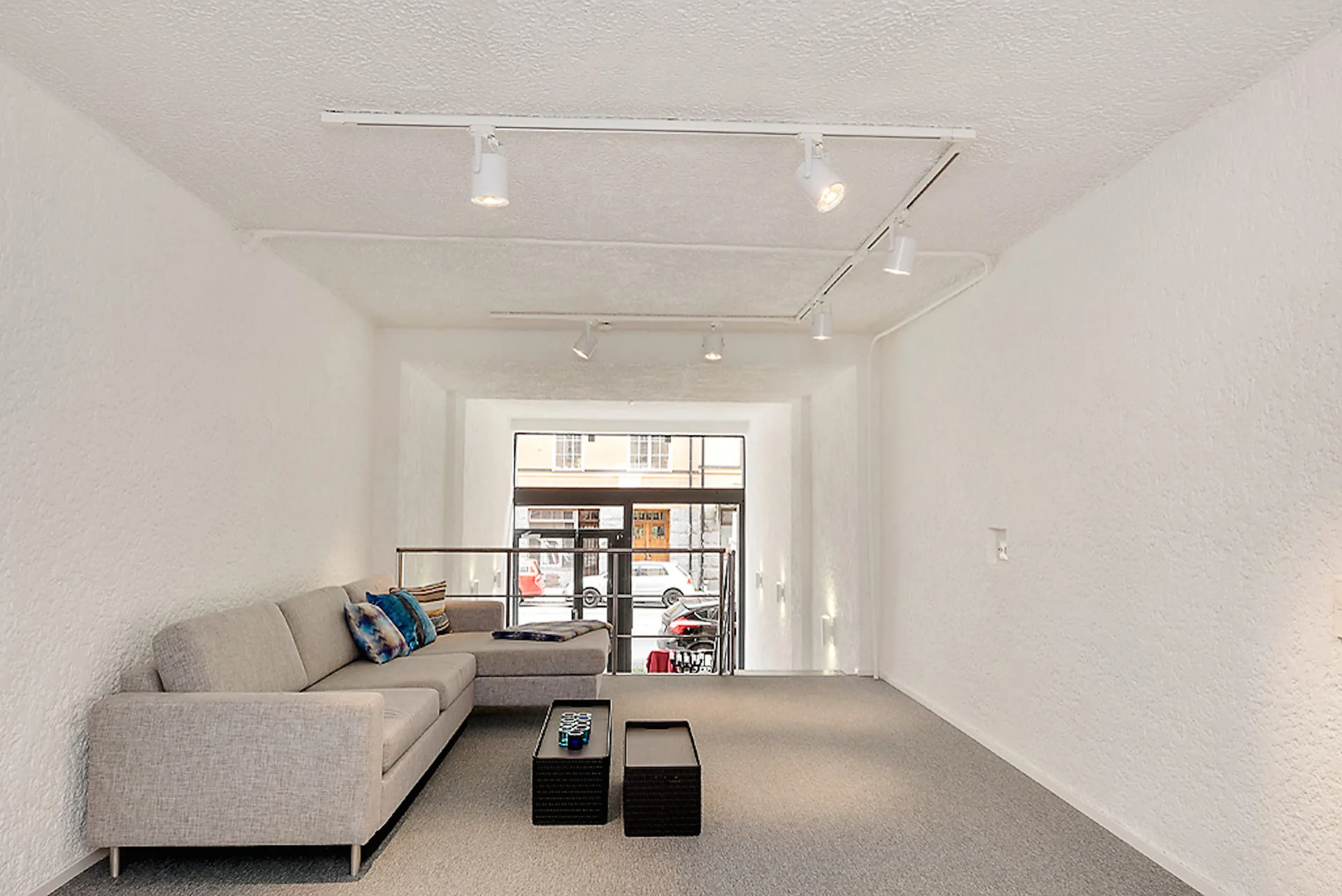 A grey sectional sofa sits on a grey carpeted floor in a loft space featuring heavily textured white walls and ceiling track lighting.