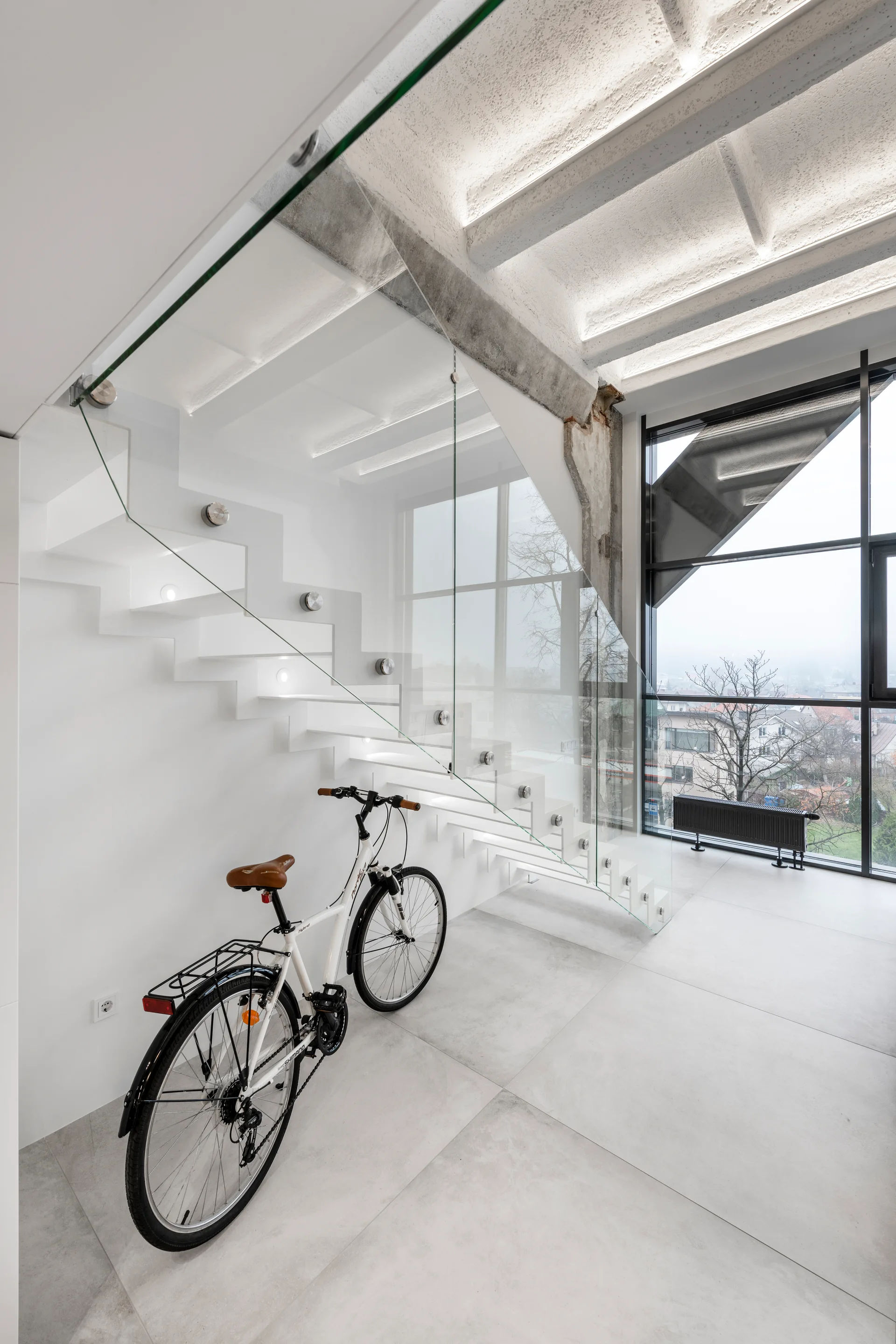 A minimalist white floating staircase with a frameless glass railing stands near large windows with a white bicycle parked on the light grey tiled floor.