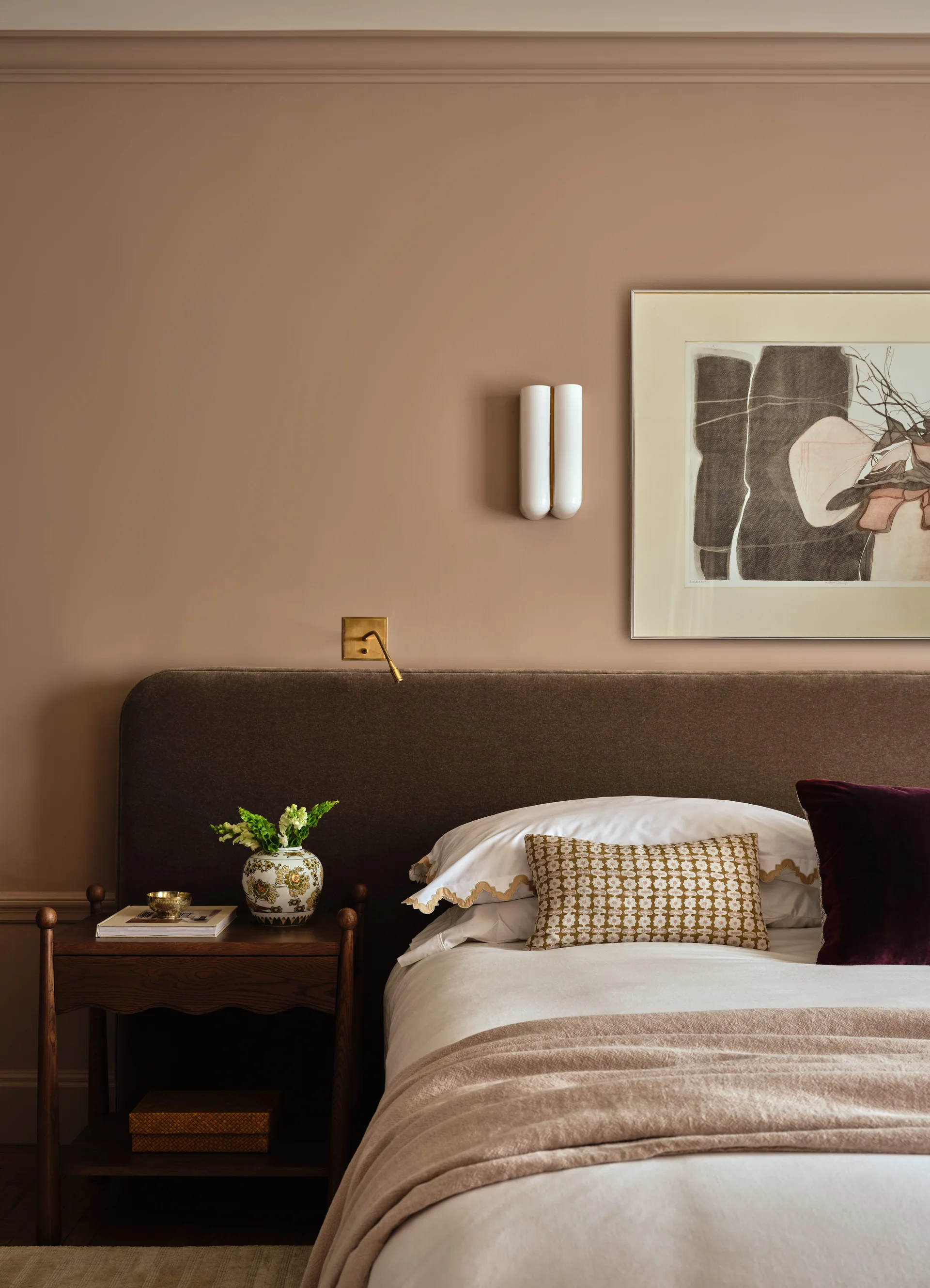 A bedroom corner features a brown upholstered headboard, a wooden nightstand, and taupe walls styled with modern sconces and a framed abstract print.
