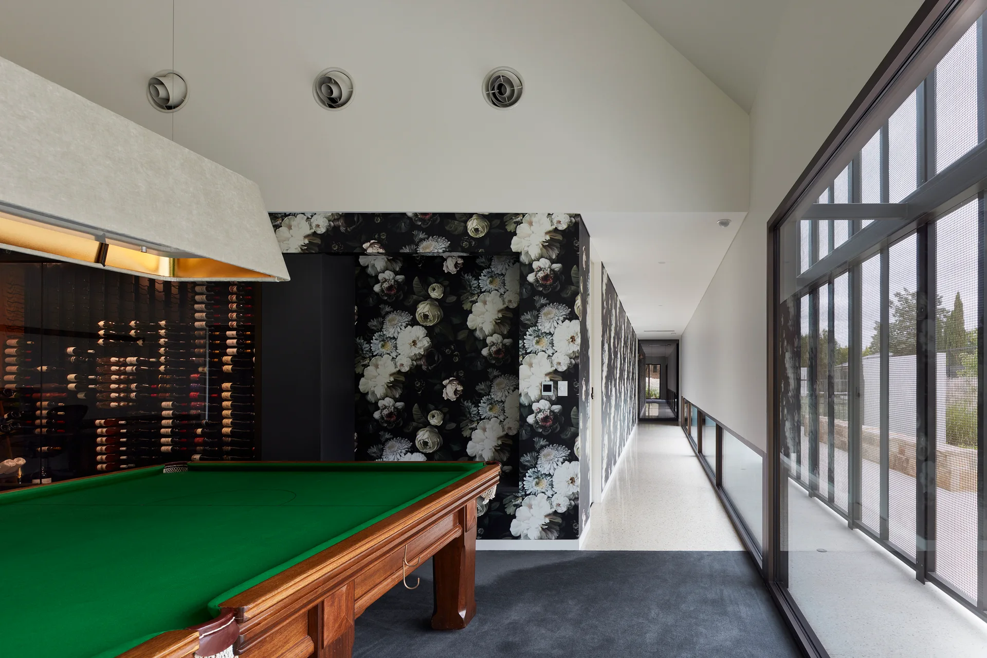 A game room with a wood pool table next to a glass wine display and a dark floral accent wall.