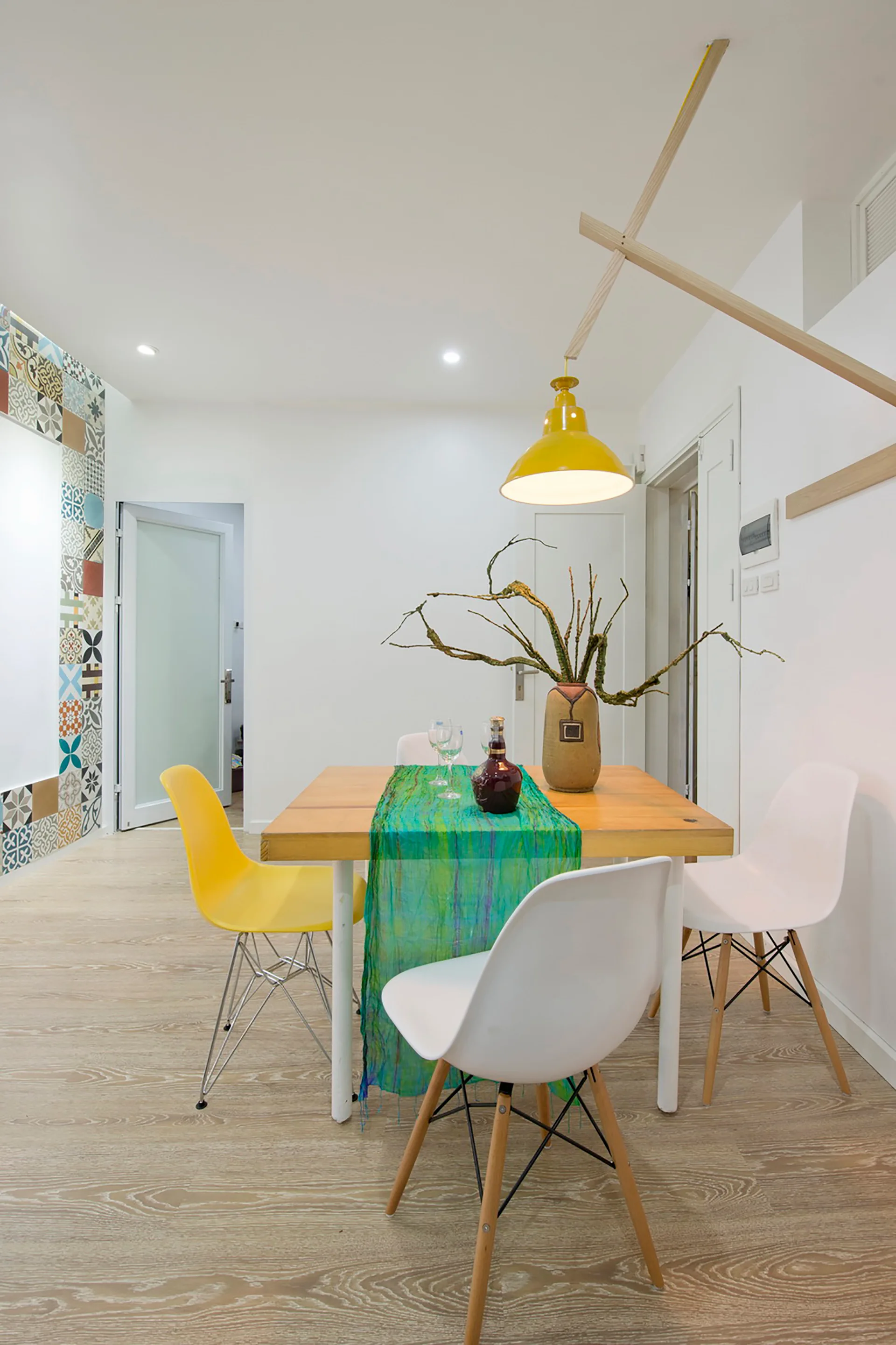 A wooden dining table with a green runner is surrounded by white and yellow chairs beneath a yellow pendant light hung from a wooden arm.