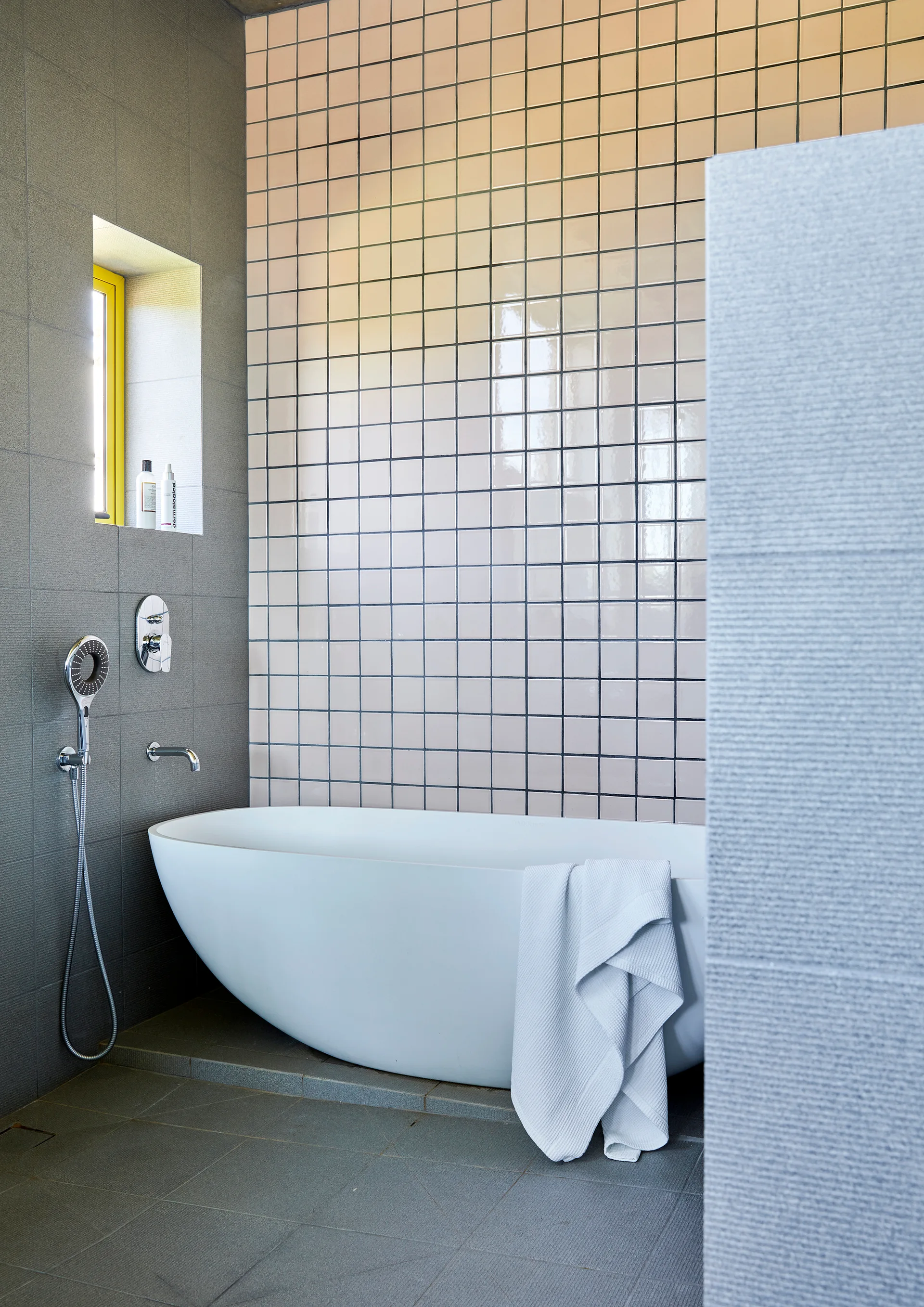 A white standalone bathtub sits against a white grid-tiled wall next to a grey wall with a small recessed window trimmed in yellow.