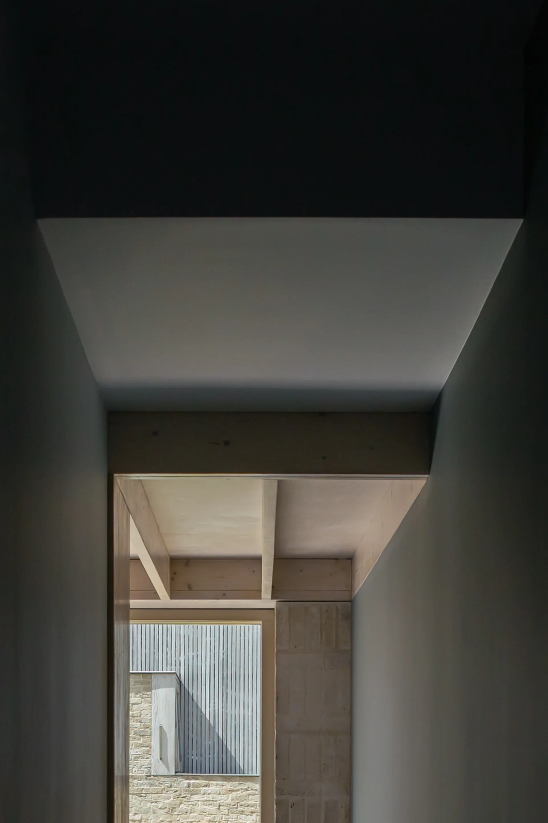 A dark hallway frames a view of a sunlit corridor featuring light wood ceiling beams and textured stone walls.