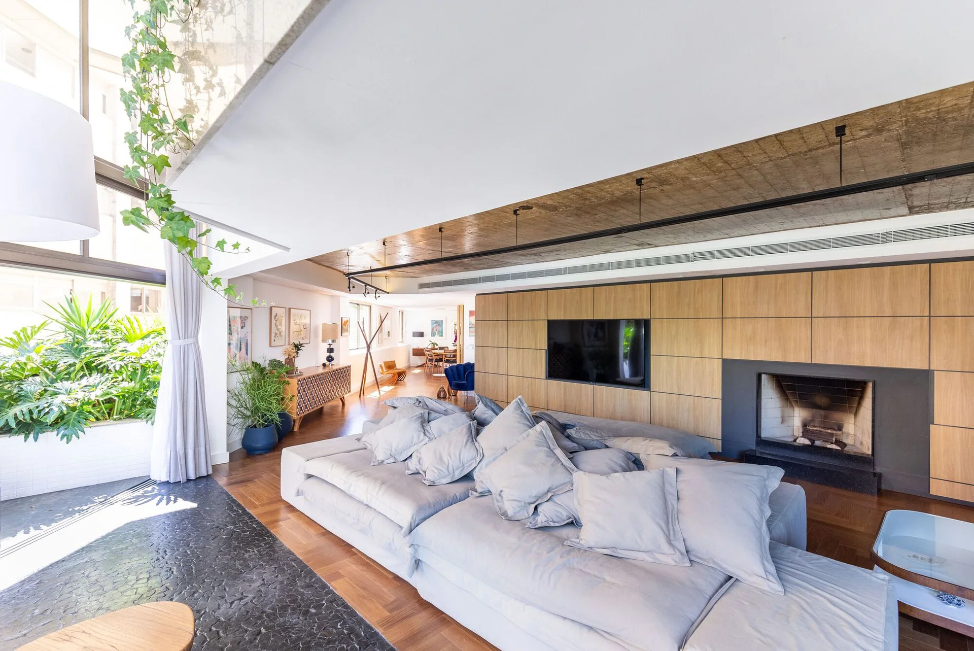 An open-plan living room features a large grey sectional sofa, a wood-paneled media wall with an integrated fireplace, and an exposed concrete ceiling.