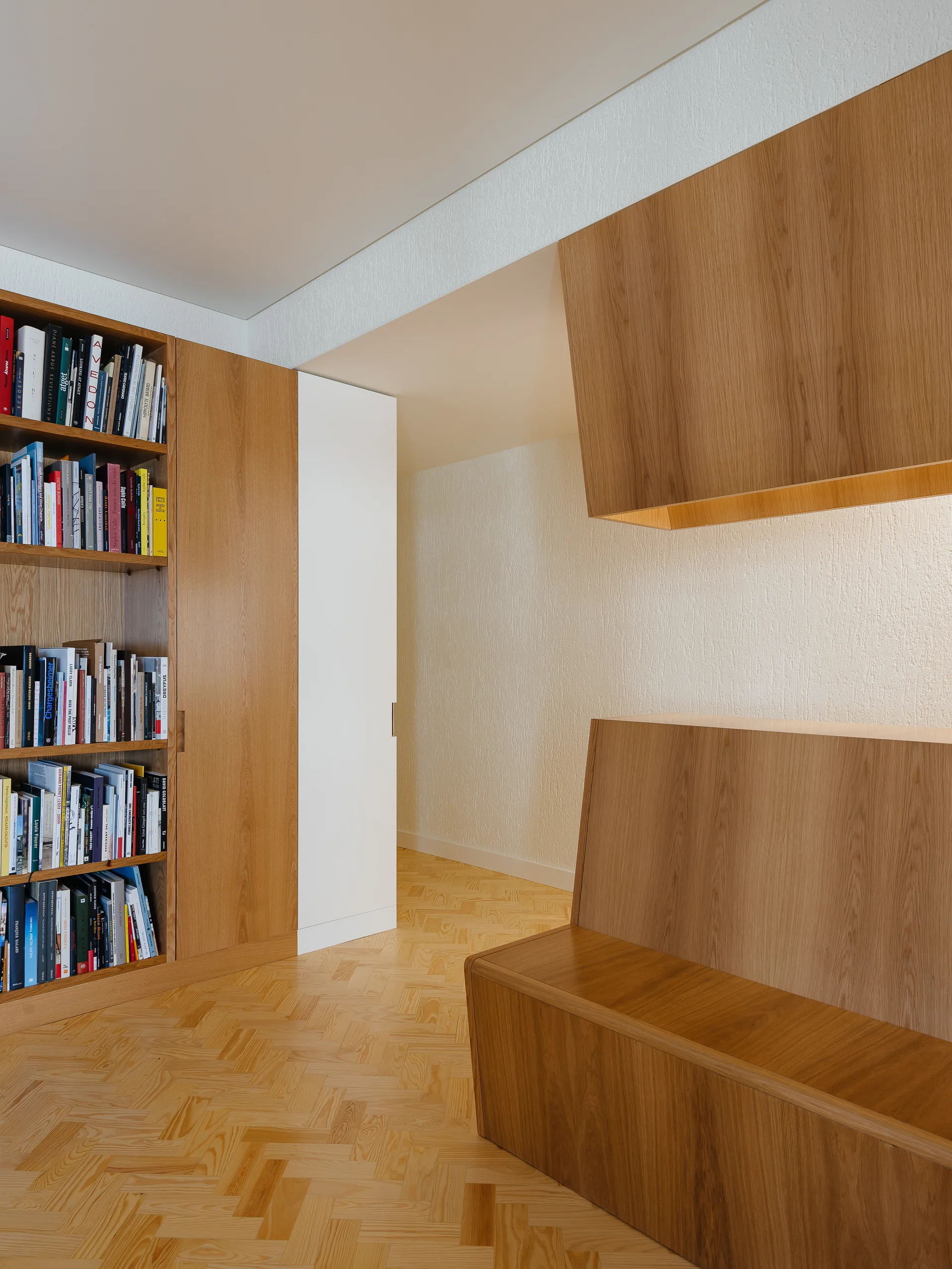 A minimalist library corner features a built-in oak bookshelf, an integrated wooden bench, textured white walls, and a herringbone parquet floor.