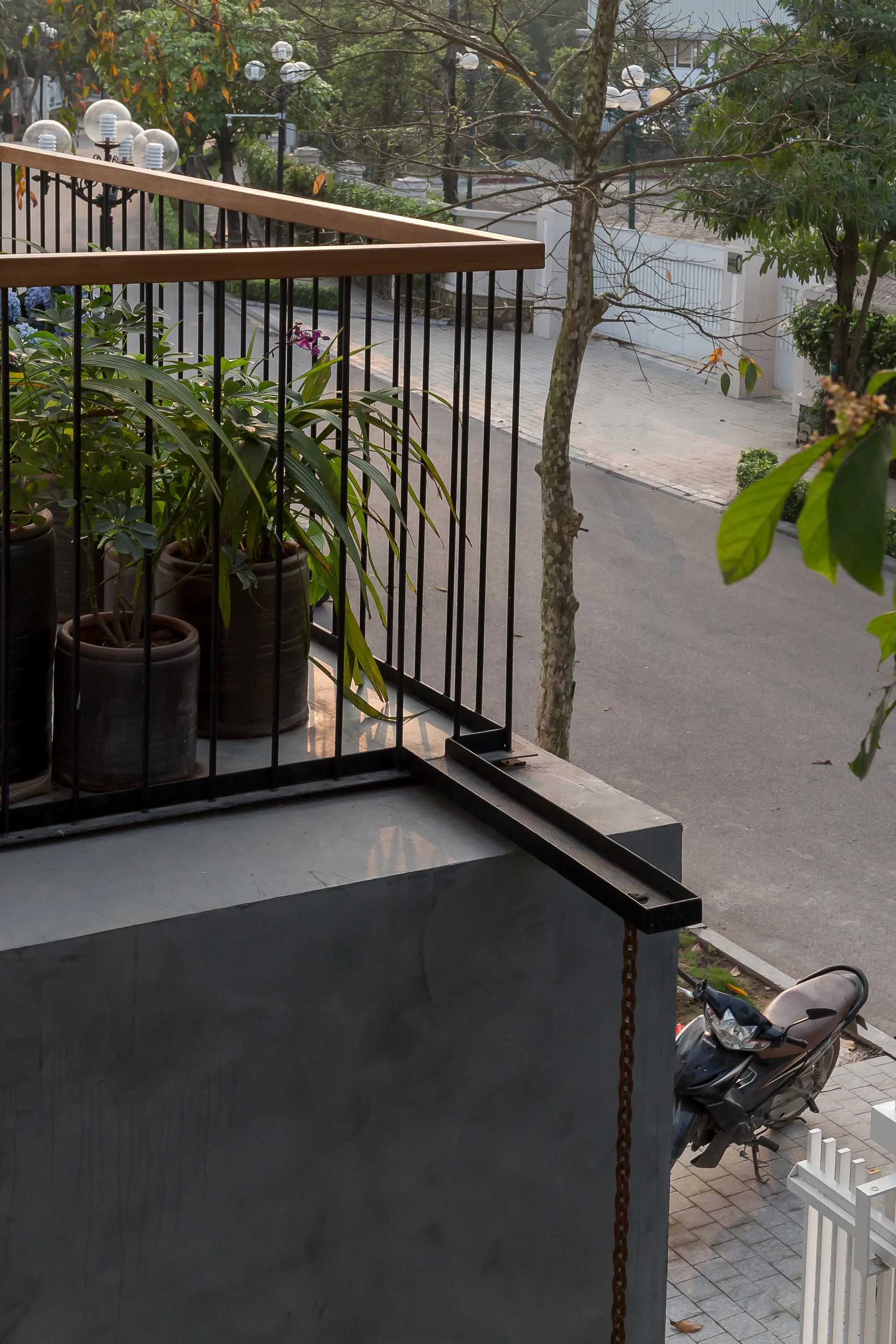 An elevated concrete balcony features a black metal railing with a wooden handrail, green potted plants, and a dark metal rain chain hanging down.