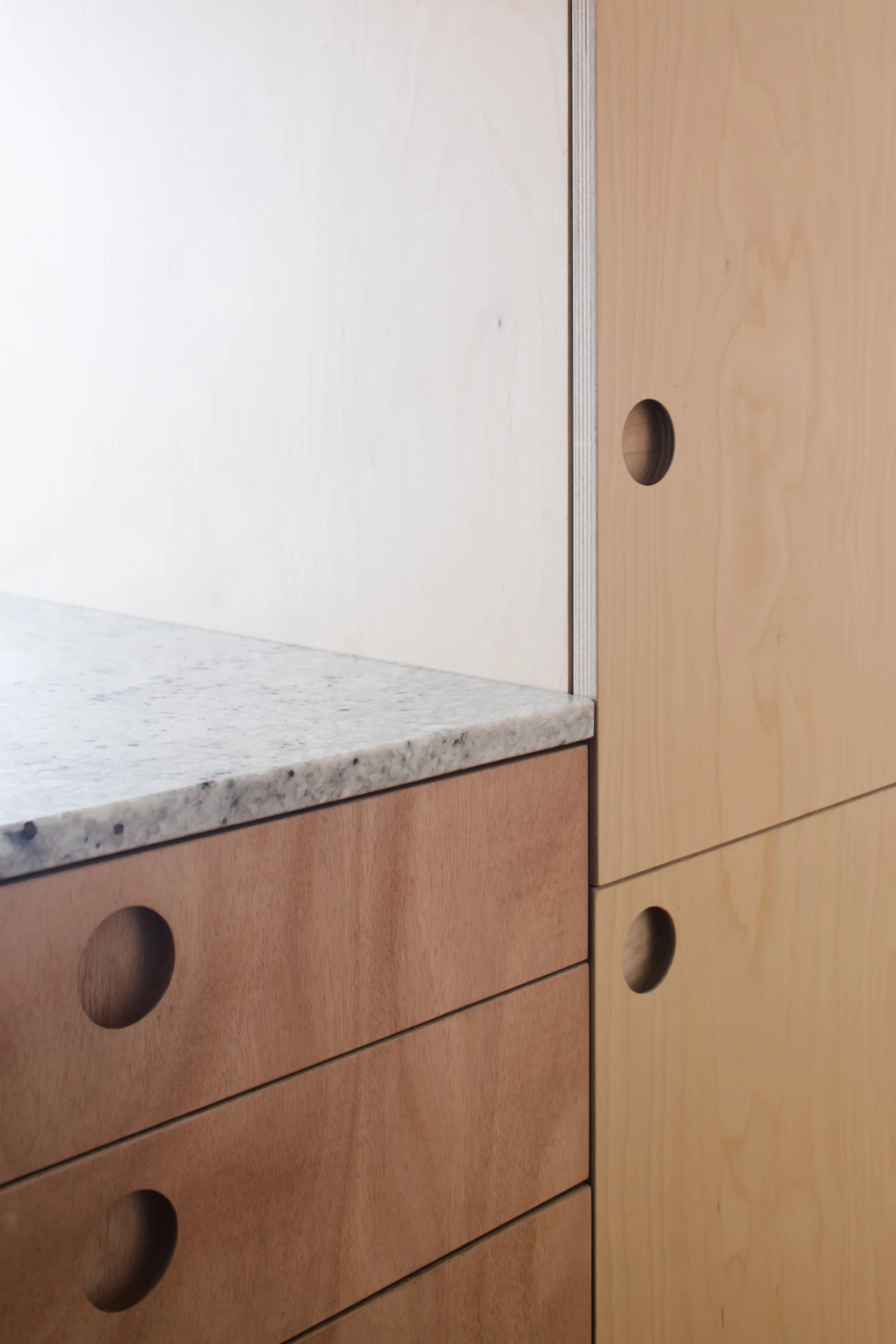 Close-up of light-colored wood plywood kitchen cabinets featuring circular cutout drawer pulls and a speckled stone countertop.