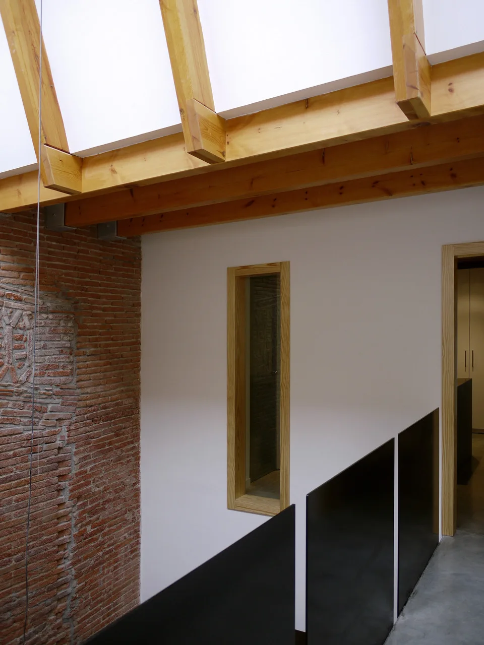 A modern hallway features exposed wooden ceiling beams, a red brick wall, a narrow vertical window, and a black panel railing.