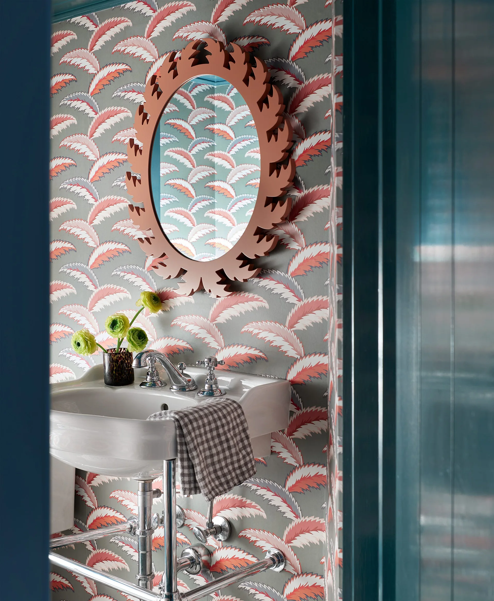 A powder room features a white console sink, patterned botanical leaf wallpaper, and a round mirror with a carved terracotta frame.